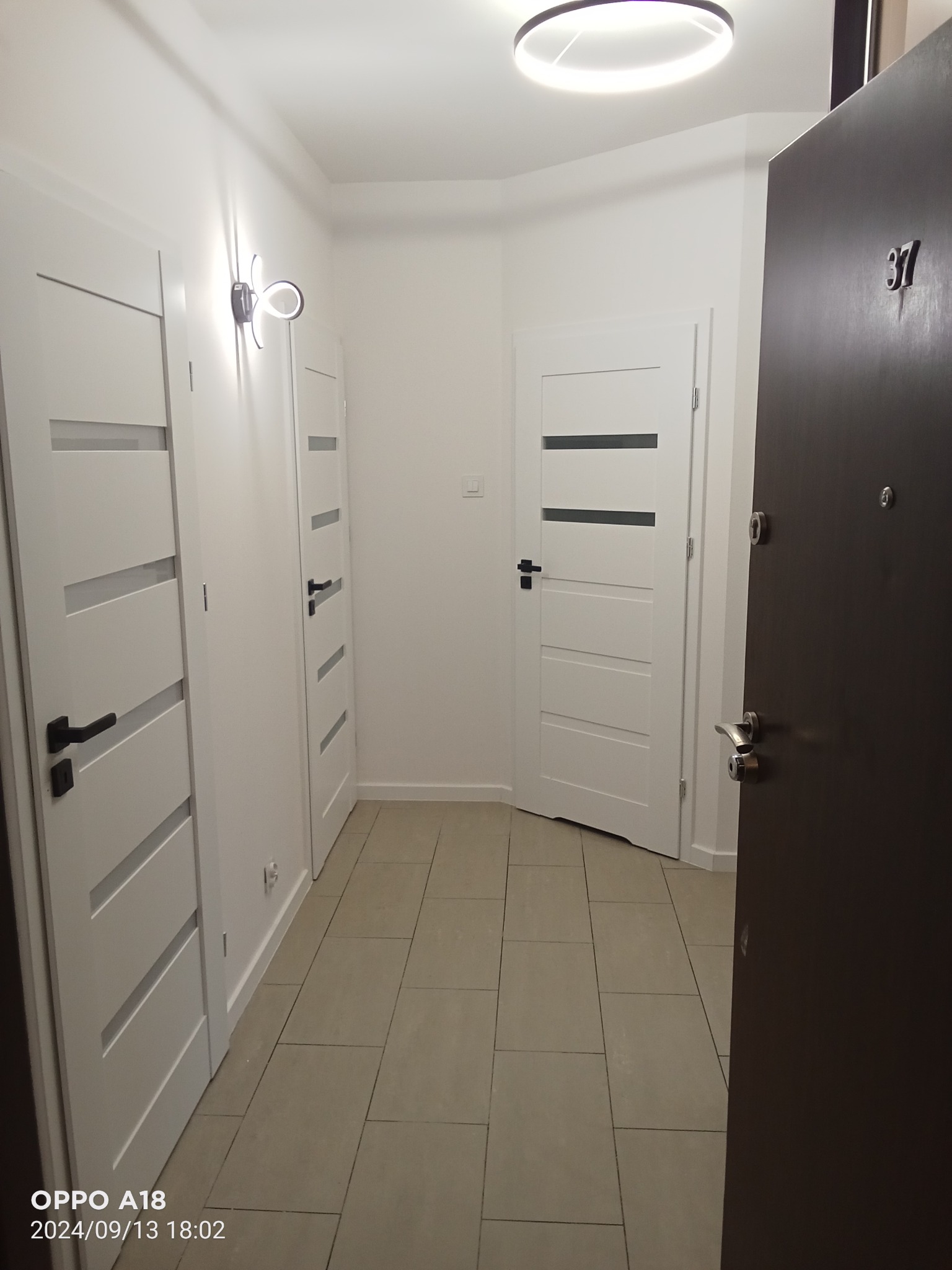 Korytarz po remoncie z trzema białymi drzwiami z poziomymi przeszkleniami, podłoga wyłożona płytkami w odcieniach beżu, na ścianie nowoczesny kinkiet, widoczny numer mieszkania 37 na ciemnych...