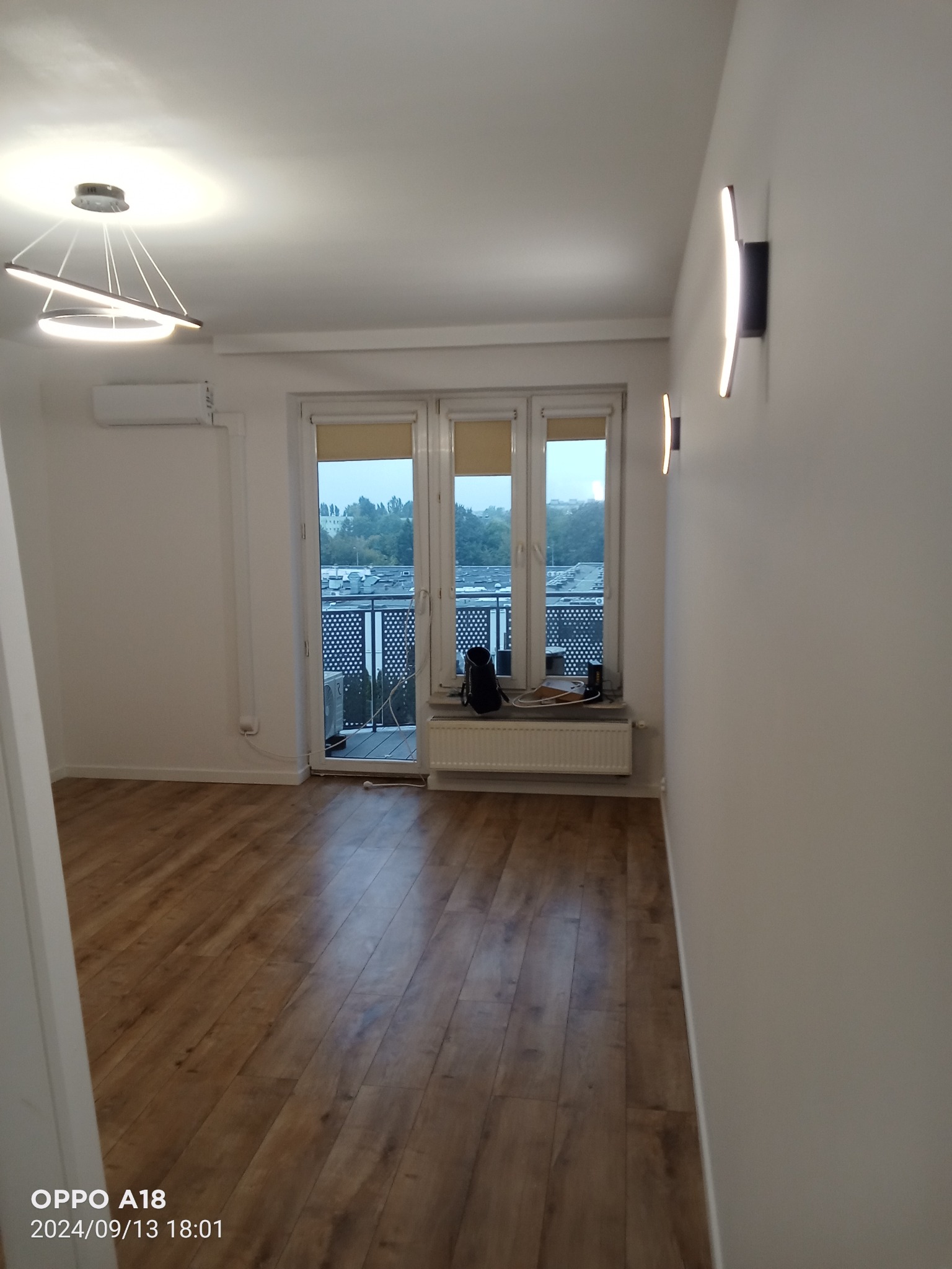 Puste, jasne wnętrze z drewnopodobną podłogą i balkonem, nowoczesne oświetlenie sufitowe i ścienne, widok na zewnątrz zza zasłoniętych okien balkonowych.