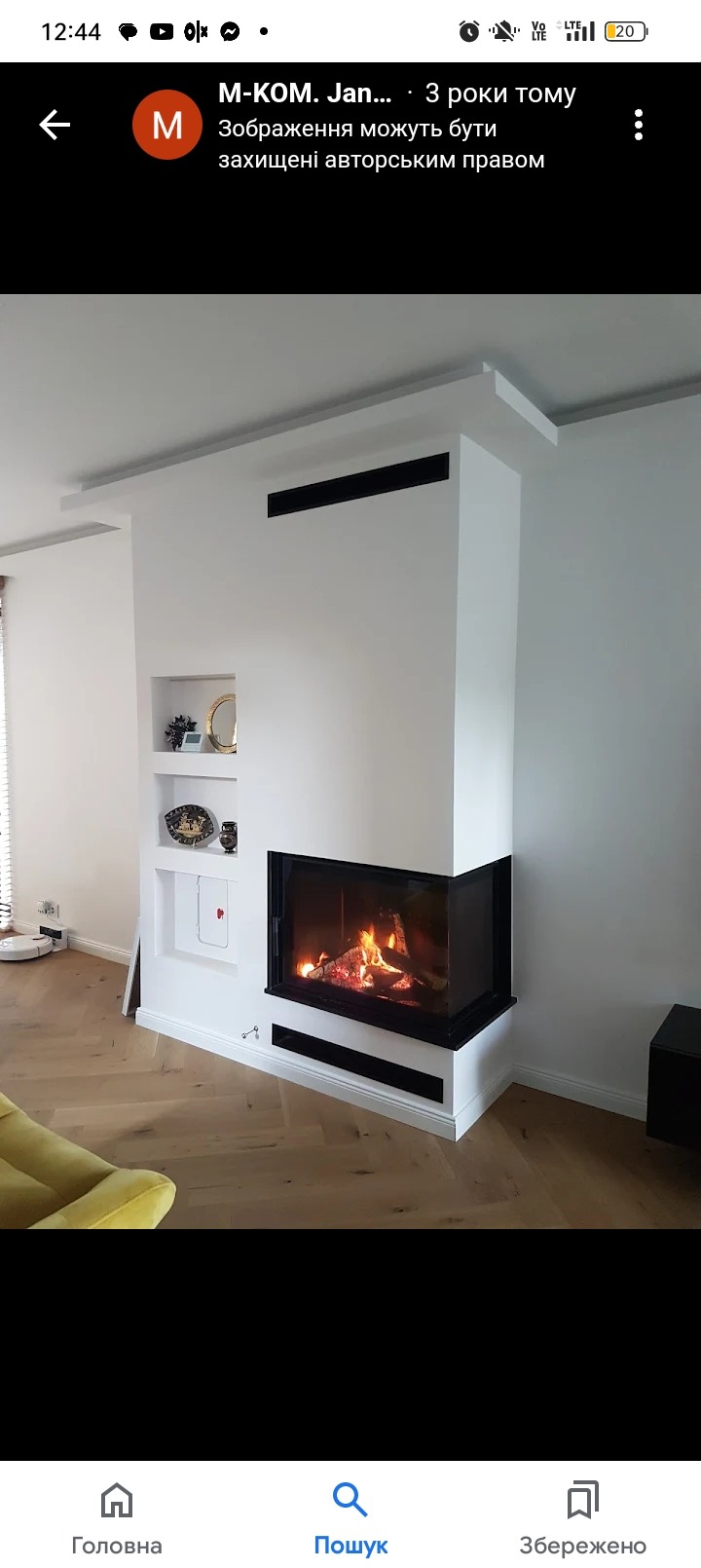 Nowoczesny kominek z czarnym obramowaniem wbudowany w białą ścianę z półkami, na podłodze parkiet ułożony w jodełkę francuską, widoczne płomienie w palenisku.