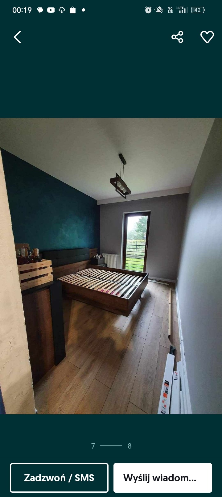 Pokój z drewnianą podłogą i ramą łóżka, ściana pomalowana na ciemny turkus, widoczna lampa sufitowa i drzwi balkonowe, listwy przypodłogowe częściowo zamontowane.