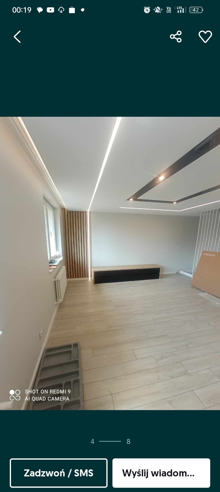 Jasne wnętrze po remoncie z drewnianą podłogą, listwami na ścianie i suficie oraz oświetleniem LED, czarna szafka RTV i karton po sprzęcie RTV.