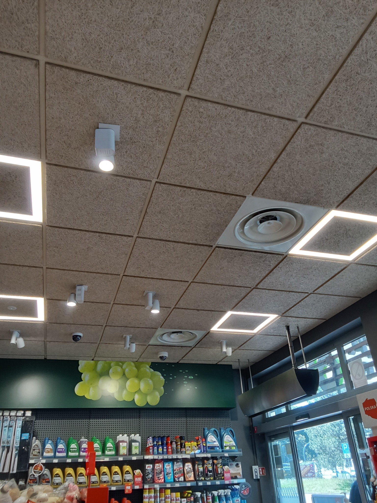 Sufit z paneli akustycznych z widocznymi oprawami oświetleniowymi LED i kratką wentylacyjną w sklepie z artykułami motoryzacyjnymi i dekoracją winogron na ścianie.