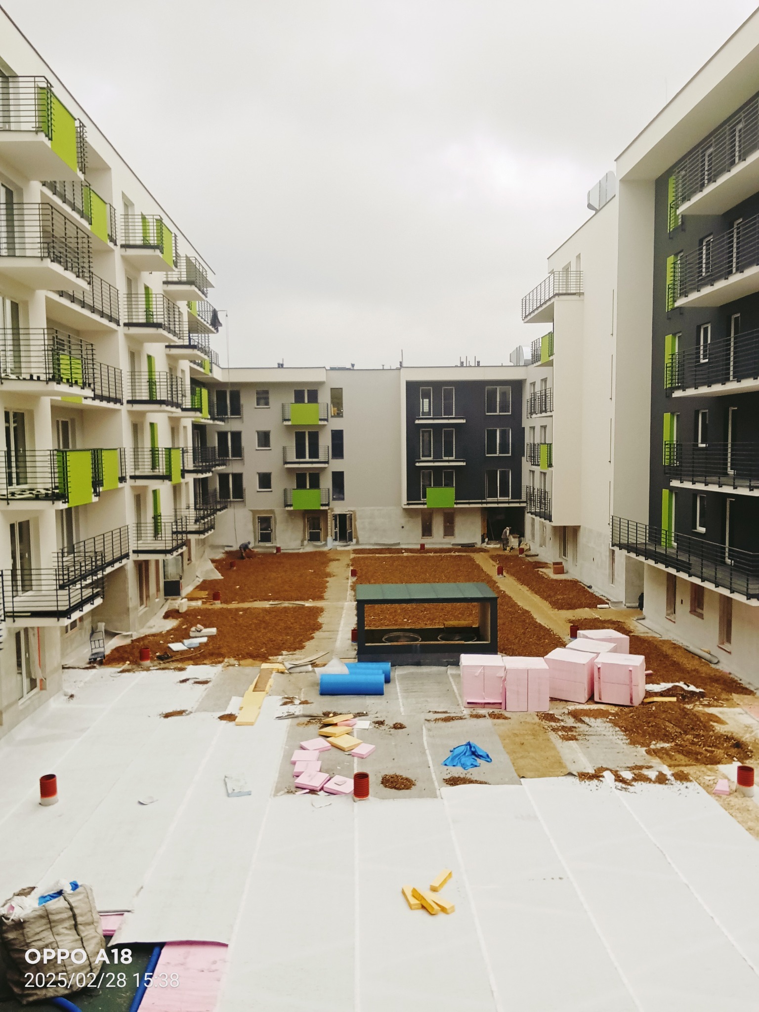 Plac budowy nowego osiedla z widocznymi blokami mieszkalnymi z balkonami w kolorze zieleni, przygotowany teren pod zieleńce oraz materiały izolacyjne na dachu, uchwycone w pochmurny dzień.