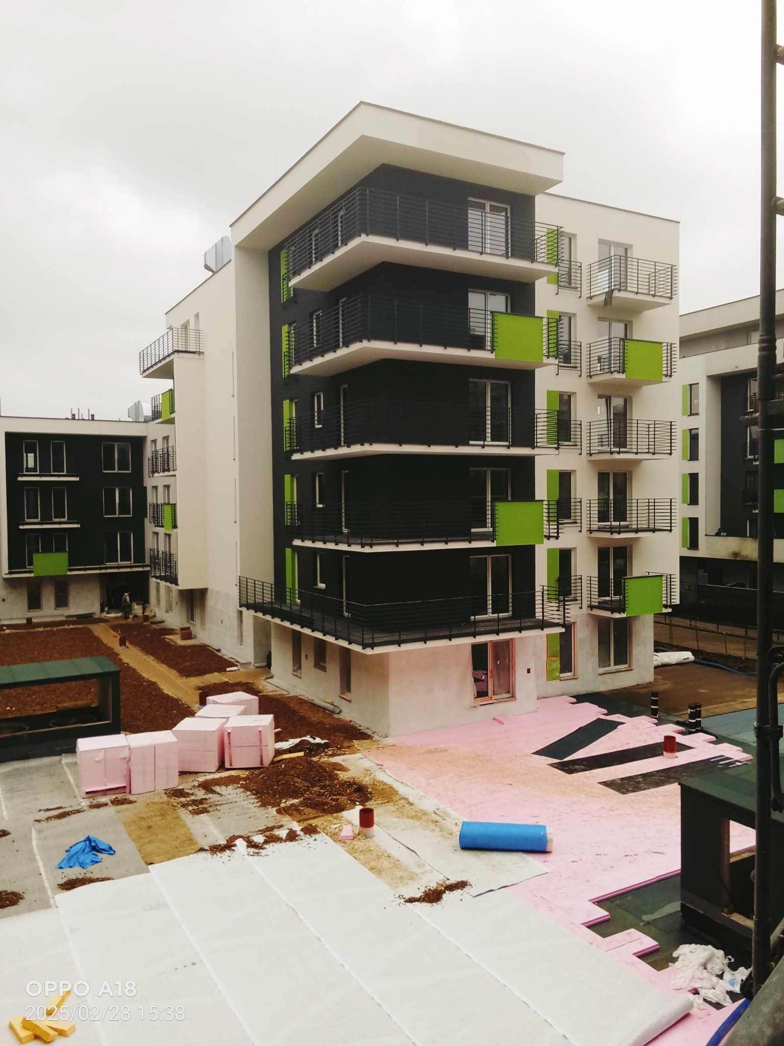 Nowoczesny budynek wielorodzinny w trakcie prac wykończeniowych, widoczna izolacja termiczna na dachu i elewacji, różowe płyty styropianowe i robotnicy w oddali.