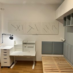 HIEX - Pokój młodzieżowy z białym biurkiem regulowanym, komodą na kółkach, łóżkiem z drewnianym stelażem i szarą tapicerowaną ścianą, oświetlony lampką biurkową i światłem z okna.