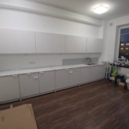 HIEX - Nowoczesna, minimalistyczna kuchnia z białymi szafkami, widok na okno z zabudową i drewnianą podłogą, częściowo zakryta kartonem na pierwszym planie.