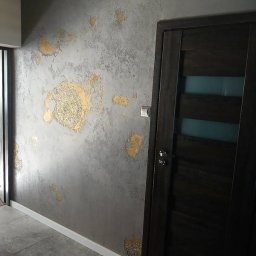 HIEX - Szara ściana z dekoracyjnym tynkiem i złotymi ornamentami, ciemne drzwi wewnętrzne z przeszkleniem, biała listwa przypodłogowa, szara podłoga z płytek.