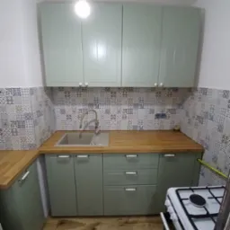 Jasnozielona zabudowa kuchenna z drewnianym blatem i mozaiką na ścianie. Ujęcie z niskiego kąta, widoczny fragment kuchenki i drewniana podłoga.