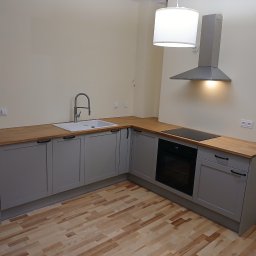 HIEX - Szara kuchnia w kształcie litery L z drewnianym blatem, okapem i lampą. Ujęcie z niskiego kąta, ukazujące drewnianą podłogę i minimalistyczny styl.