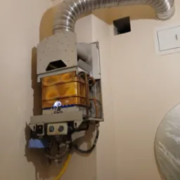 Działający gazowy piec dwufunkcyjny z widocznym płomieniem, podłączony do komina elastyczną rurą. Na ścianie widoczne elementy instalacji.