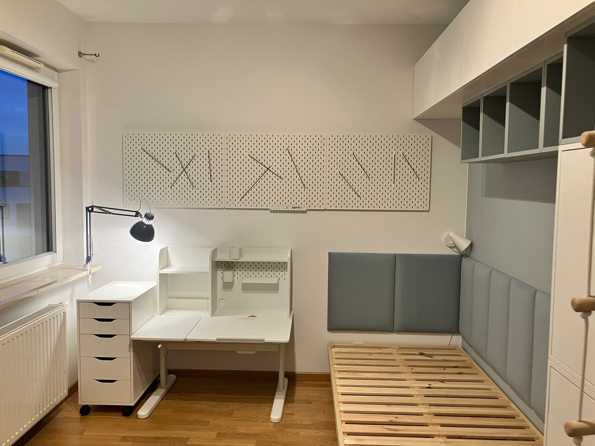 Pokój młodzieżowy z białym biurkiem regulowanym, komodą na kółkach, łóżkiem z drewnianym stelażem i szarą tapicerowaną ścianą, oświetlony lampką biurkową i światłem z okna.