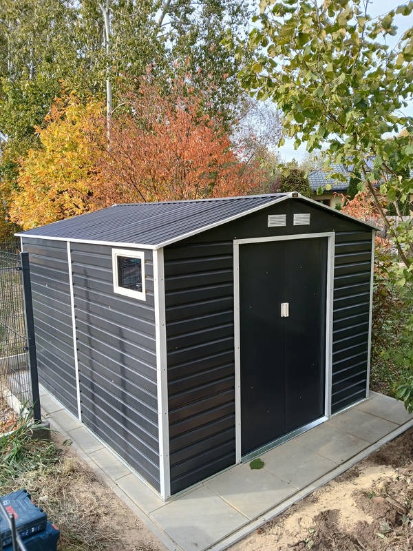 Metalowy domek narzędziowy z oknem, postawiony na betonowej wylewce w Nadarzynie, na tle jesiennych drzew.