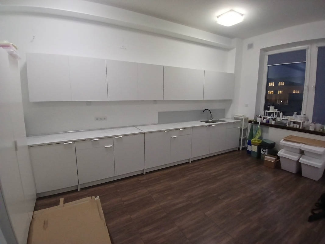 Nowoczesna, minimalistyczna kuchnia z białymi szafkami, widok na okno z zabudową i drewnianą podłogą, częściowo zakryta kartonem na pierwszym planie.