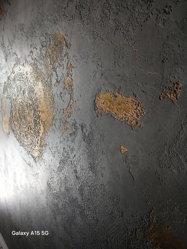 Dekoracyjna ściana z szarą, chropowatą fakturą i złotymi akcentami, w tym okrągły ornament w lewym górnym rogu.