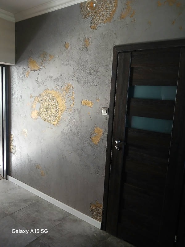 Szara ściana z dekoracyjnym tynkiem i złotymi ornamentami, ciemne drzwi wewnętrzne z przeszkleniem, biała listwa przypodłogowa, szara podłoga z płytek.