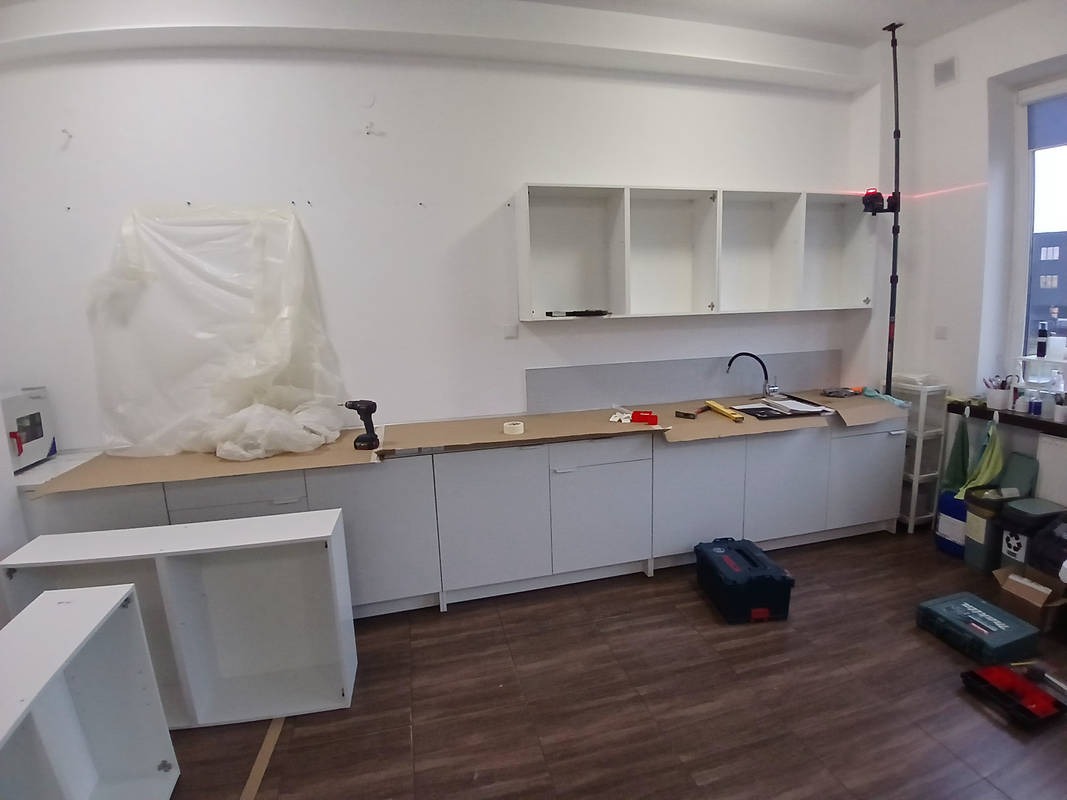 Montaż białych szafek kuchennych: dolne szafki zamontowane, blat roboczy z kartonową osłoną, na blacie poziomica i laser krzyżowy, górne szafki wiszące, na podłodze skrzynki z narzędziami.