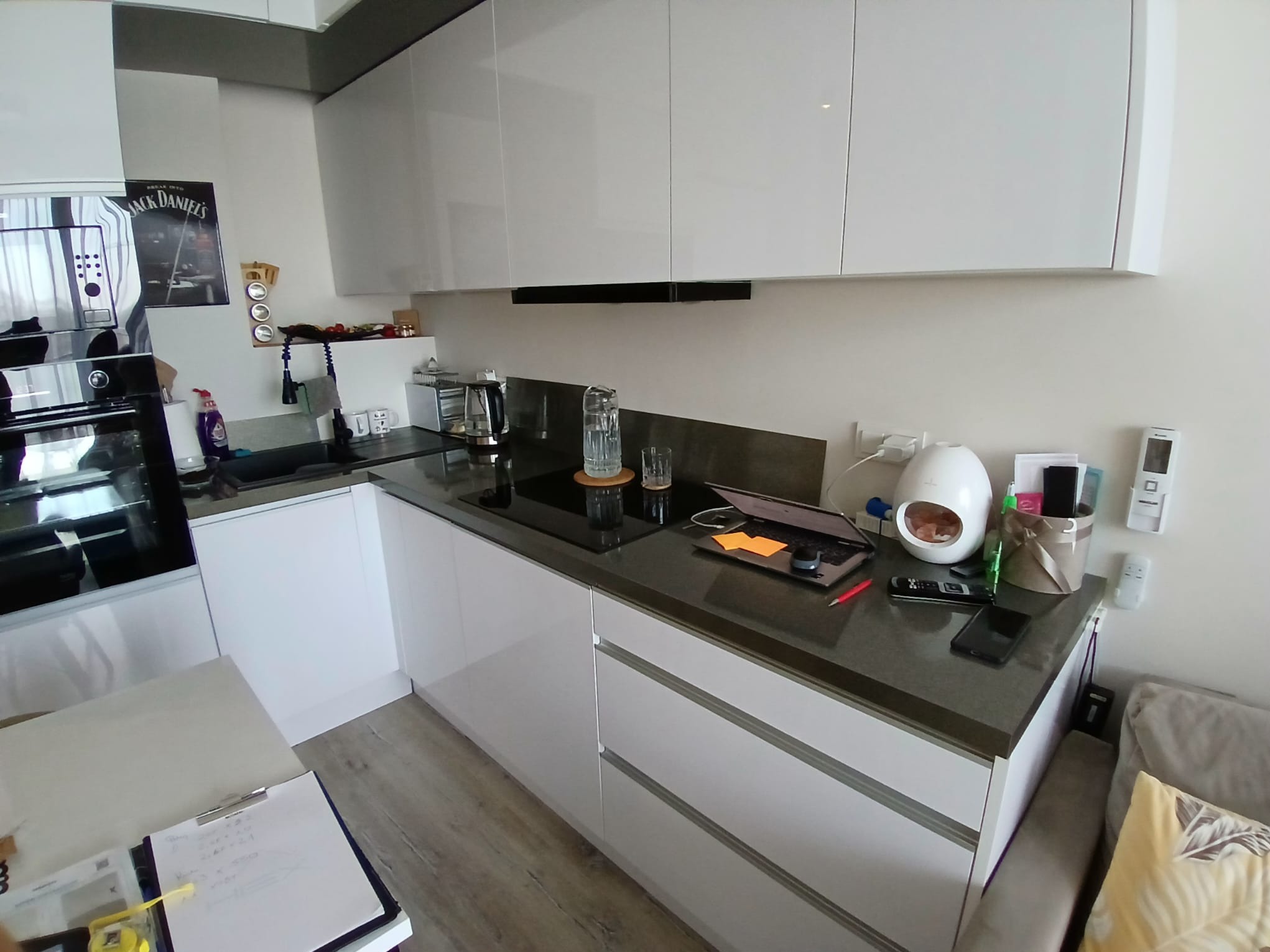 Nowoczesna kuchnia z białymi szafkami, czarnym blatem, płytą indukcyjną, laptopem i elementami dekoracyjnymi na blacie, widoczne notatki na stole.