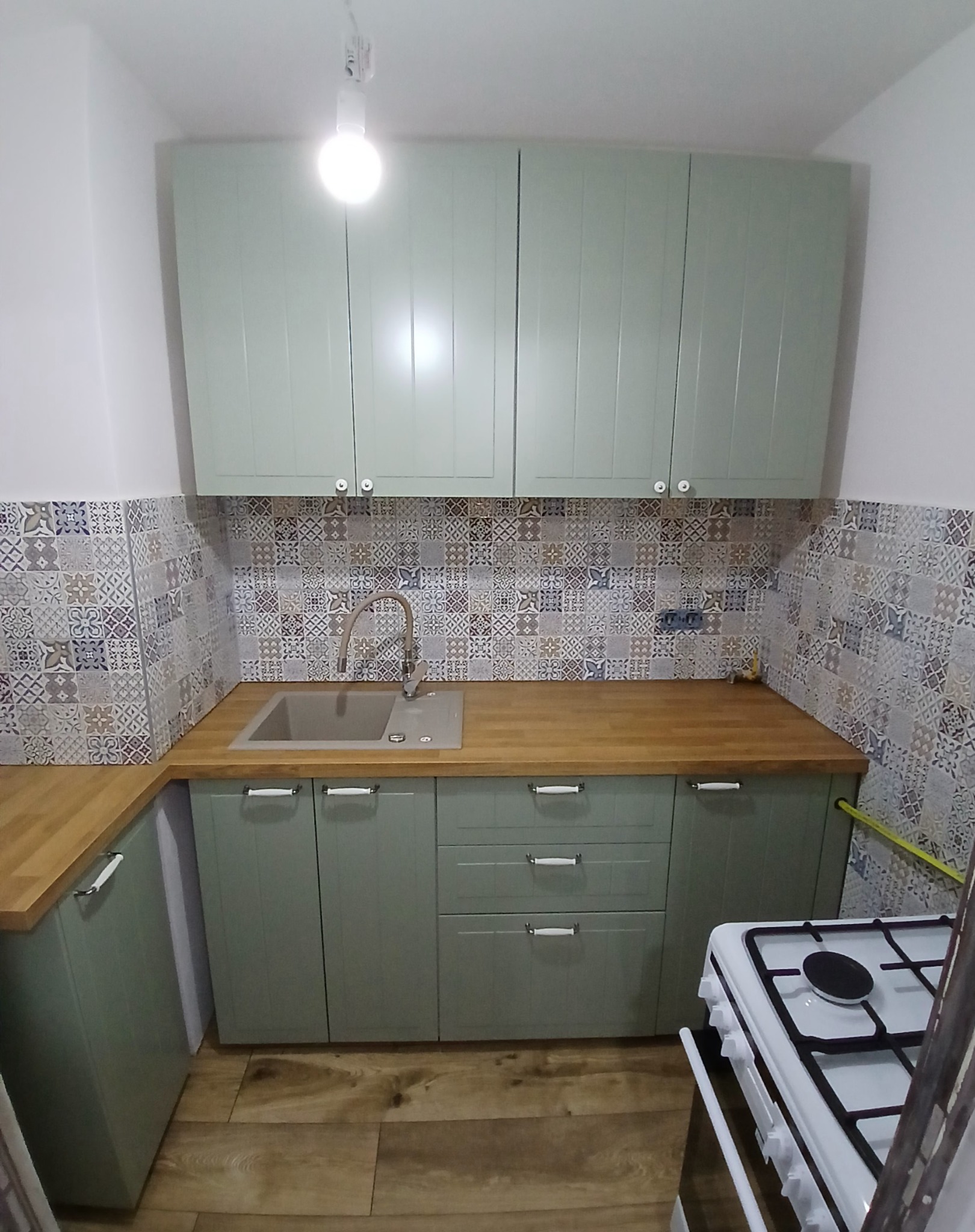 Jasnozielona zabudowa kuchenna z drewnianym blatem i mozaiką na ścianie. Ujęcie z niskiego kąta, widoczny fragment kuchenki i drewniana podłoga.