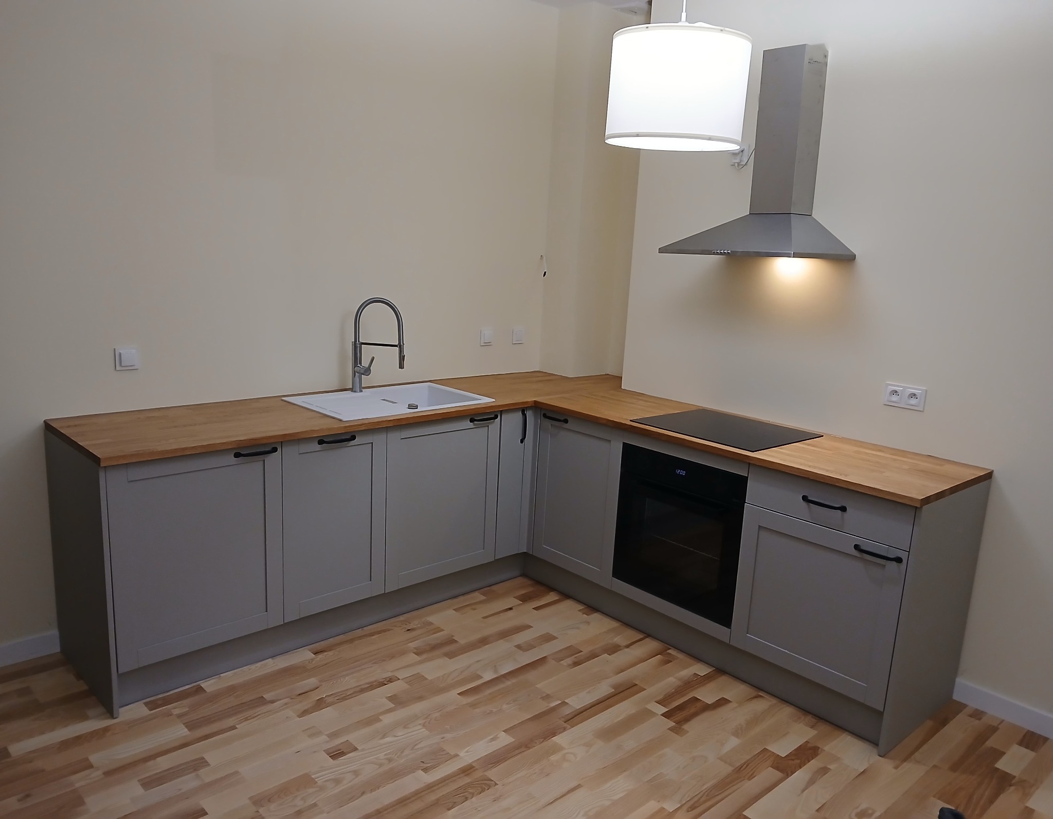 Szara kuchnia w kształcie litery L z drewnianym blatem, okapem i lampą. Ujęcie z niskiego kąta, ukazujące drewnianą podłogę i minimalistyczny styl.