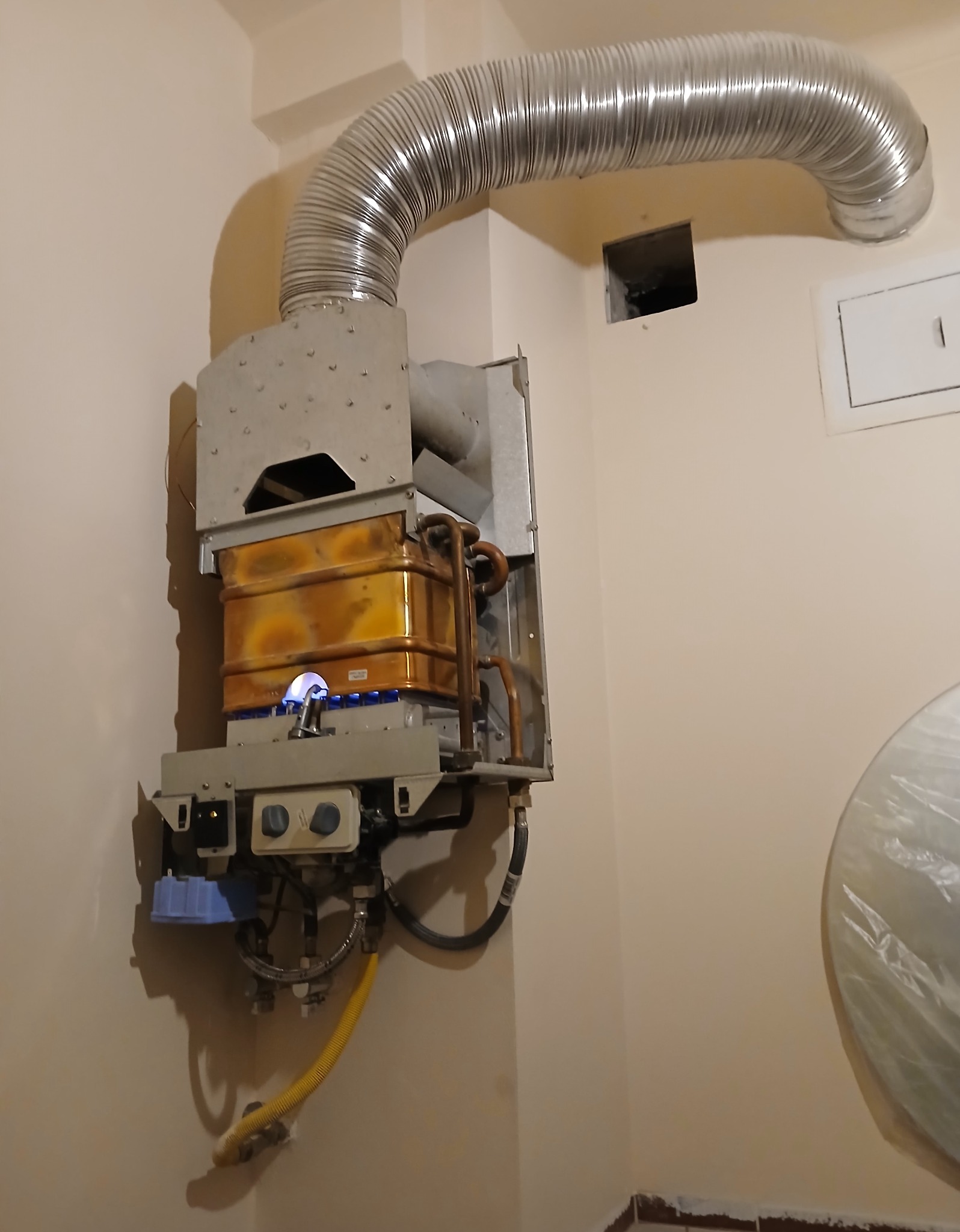 Działający gazowy piec dwufunkcyjny z widocznym płomieniem, podłączony do komina elastyczną rurą. Na ścianie widoczne elementy instalacji.