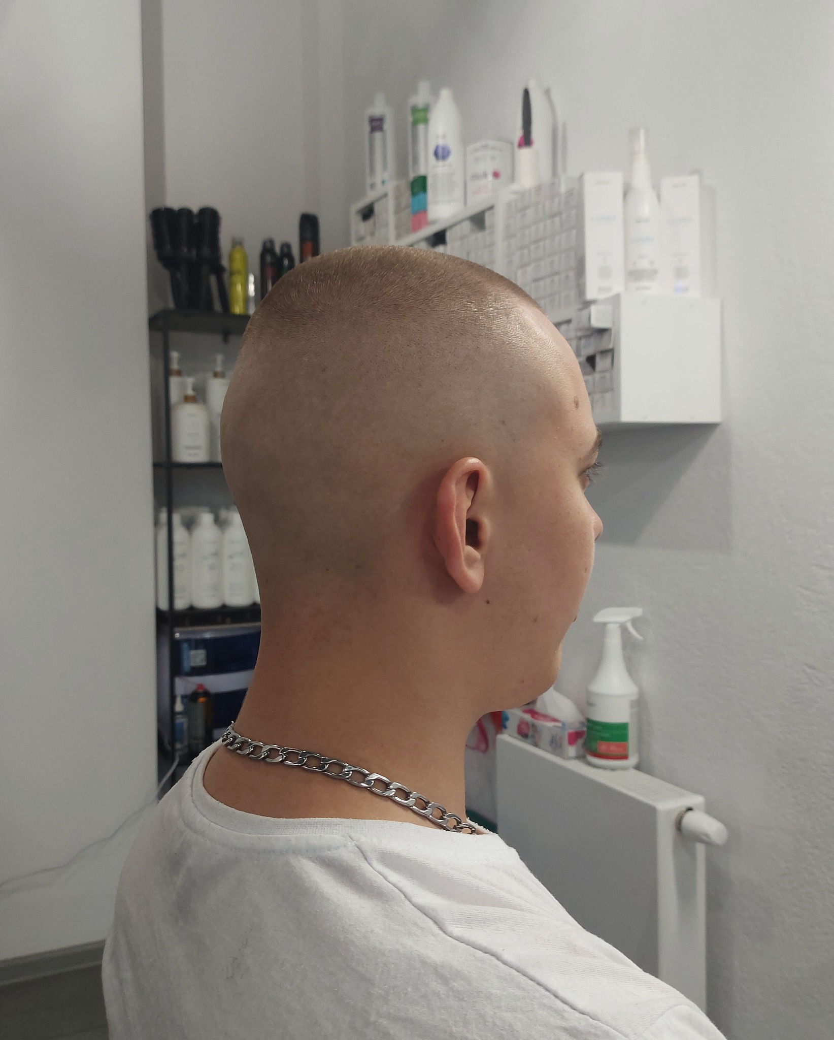 Mężczyzna z bardzo krótko ostrzyżonymi włosami, widok z profilu, srebrny łańcuszek na szyi, w tle półki z kosmetykami do włosów.