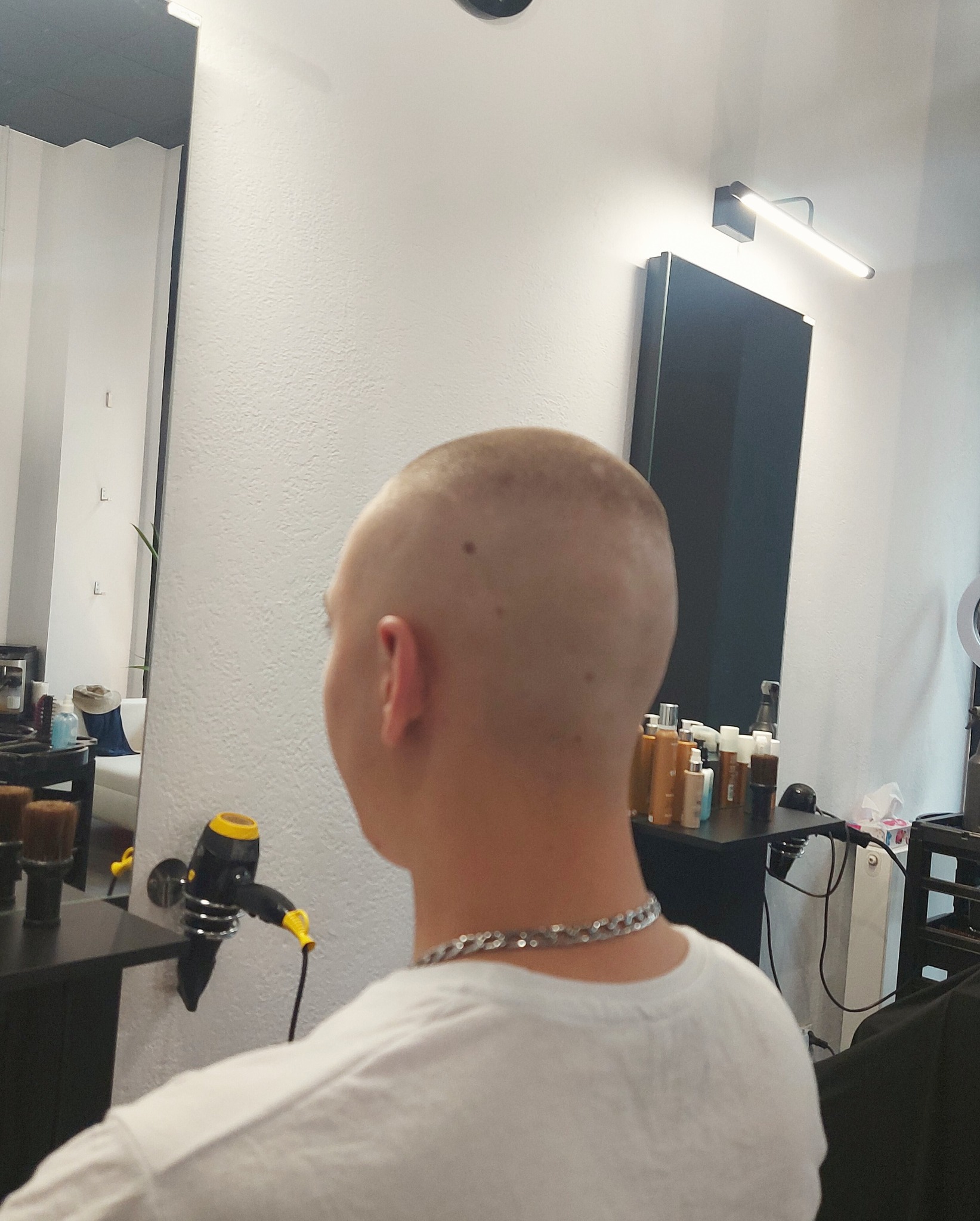 Mężczyzna z ogoloną głową, widok z tyłu, w salonie, z widocznym suszarką i kosmetykami na półce.