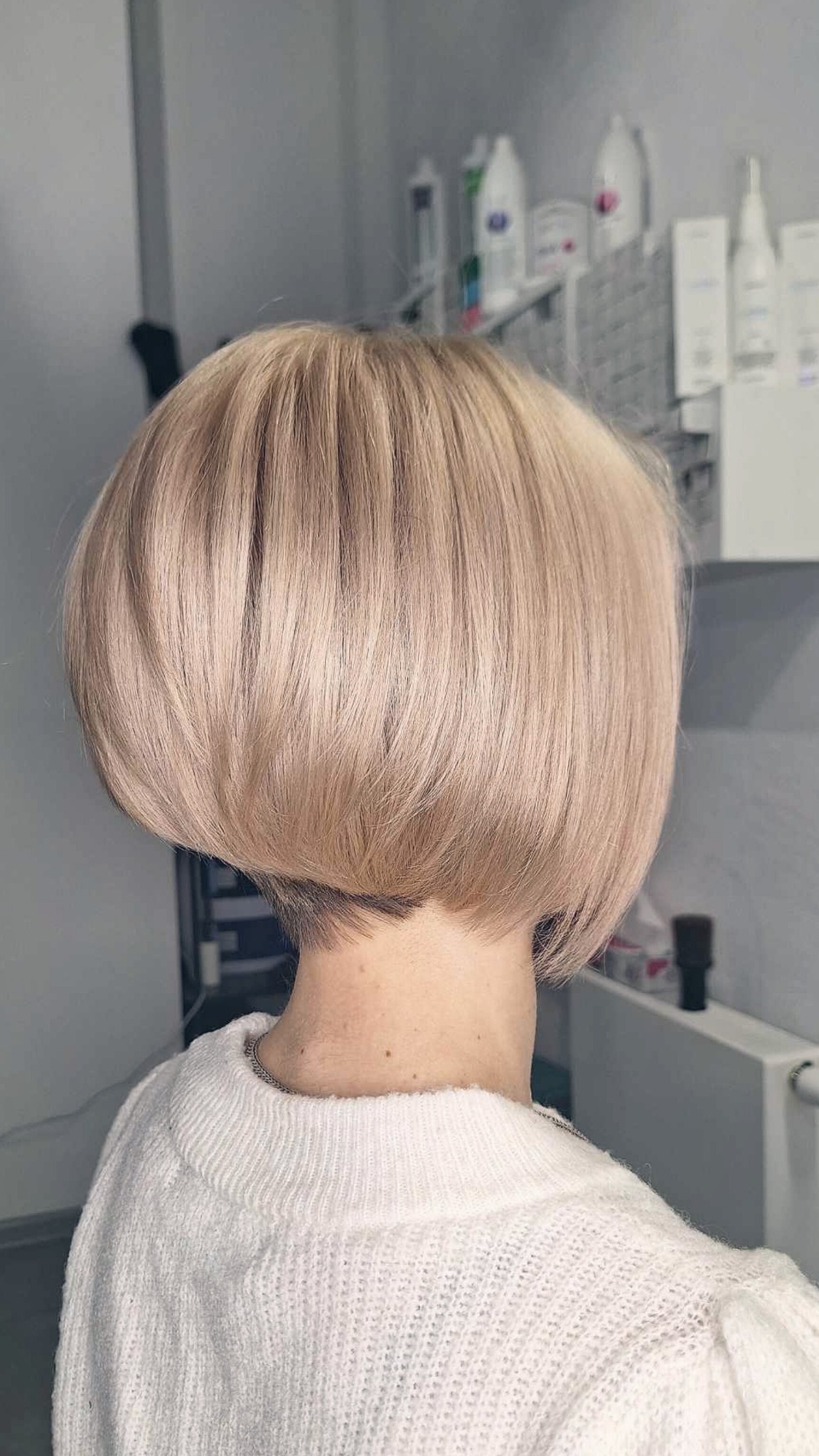 Jasny blond bob z delikatnym podcięciem, widok z tyłu, na tle półek z produktami fryzjerskimi.