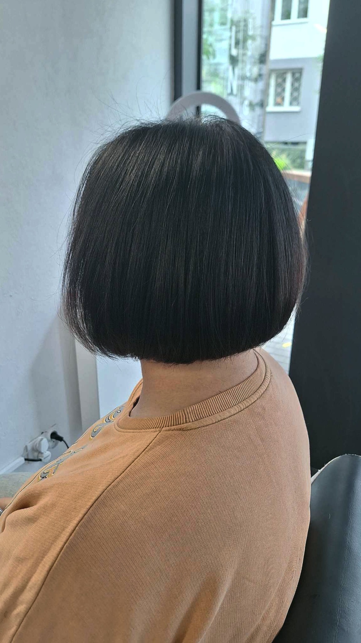 Krótkie, ciemne włosy obcięte w bob z widocznymi delikatnymi refleksami, widok z tyłu, osoba w beżowej bluzie.