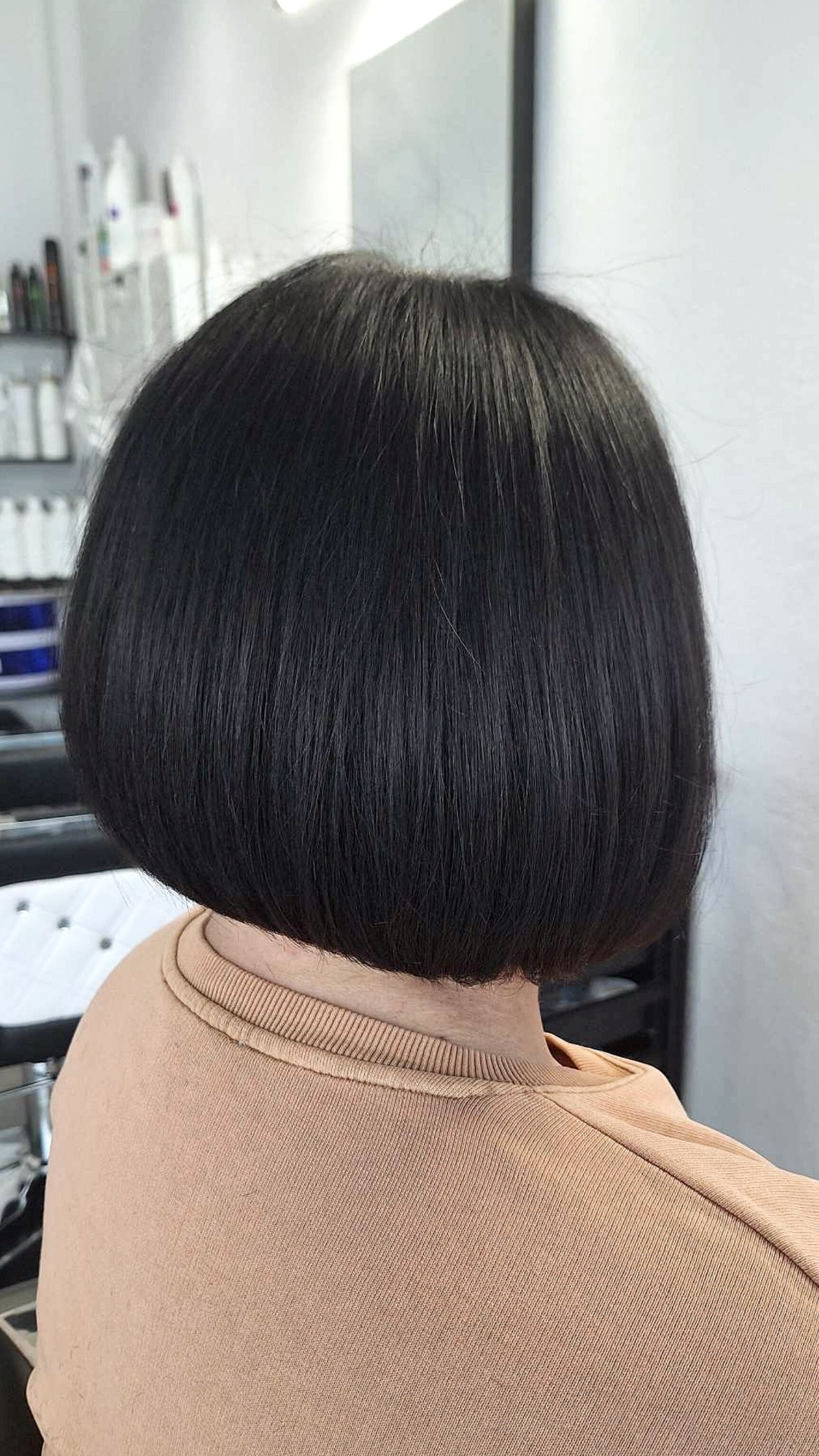 Krótkie, czarne, lśniące włosy ułożone w bob z widoczną linią cięcia na karku, widok z tyłu, w tle kosmetyki na półce.