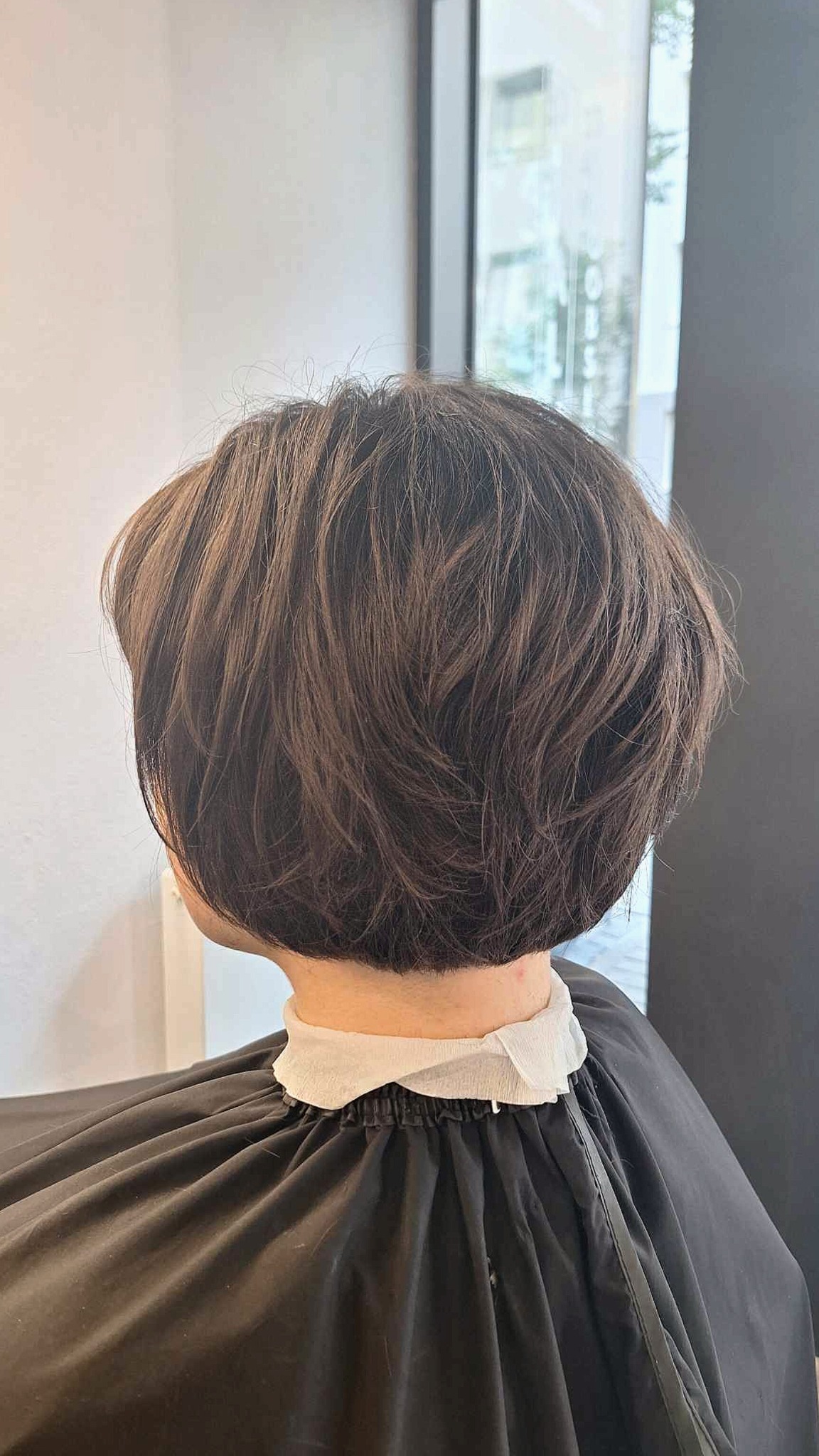 Krótkie, brązowe włosy po strzyżeniu bob, widok z tyłu, z założonym czarnym fartuchem ochronnym z białym kołnierzem.