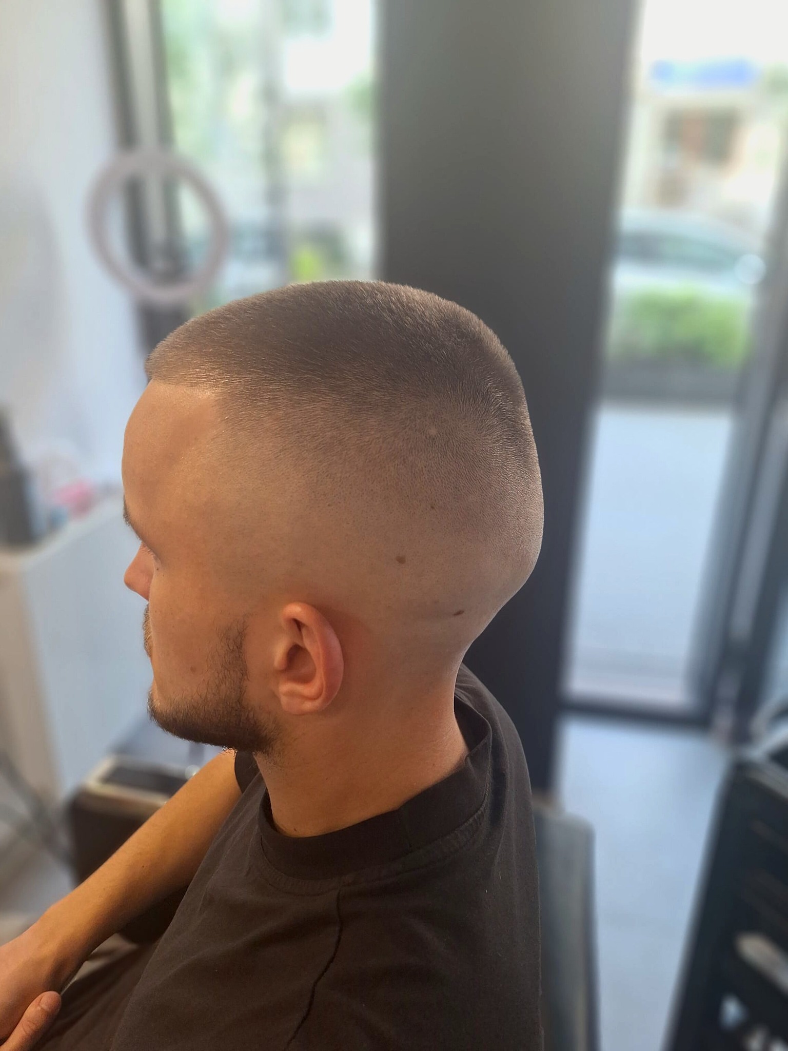 Męska fryzura z bardzo krótkimi włosami, widok z profilu, z delikatnym zarostem, w tle rozmyte wnętrze salonu.