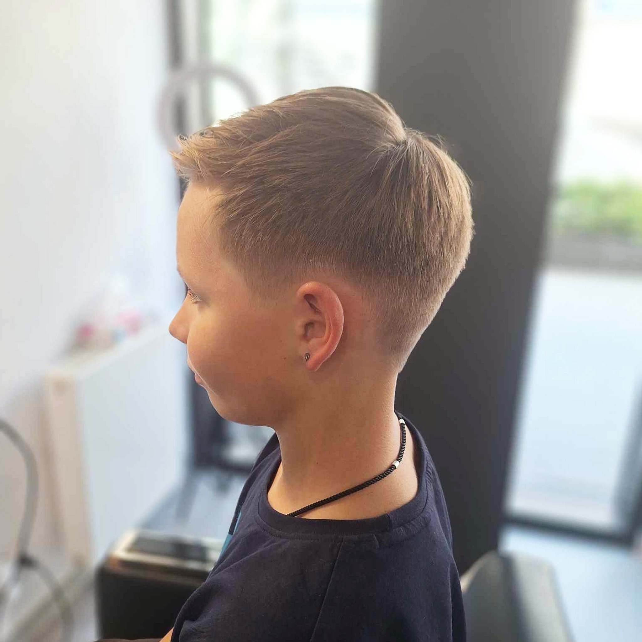 Chłopiec z modną, krótko ostrzyżoną fryzurą, widok z profilu, z delikatnym kolczykiem w uchu i czarnym naszyjnikiem z białymi akcentami.