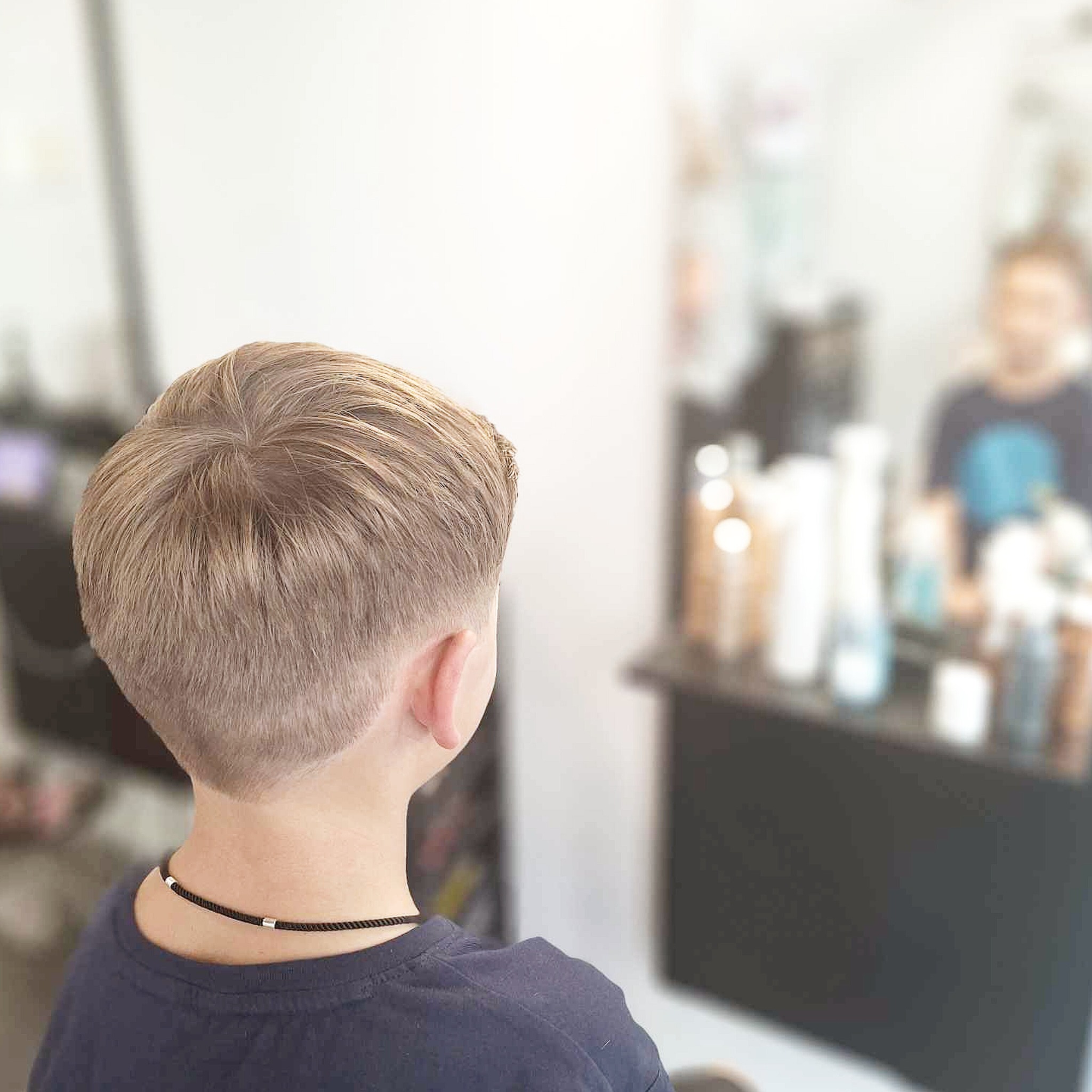 Chłopięca fryzura z blondu z widocznym gradientem, widok z tyłu, w tle rozmyte lustro i półka z kosmetykami fryzjerskimi.