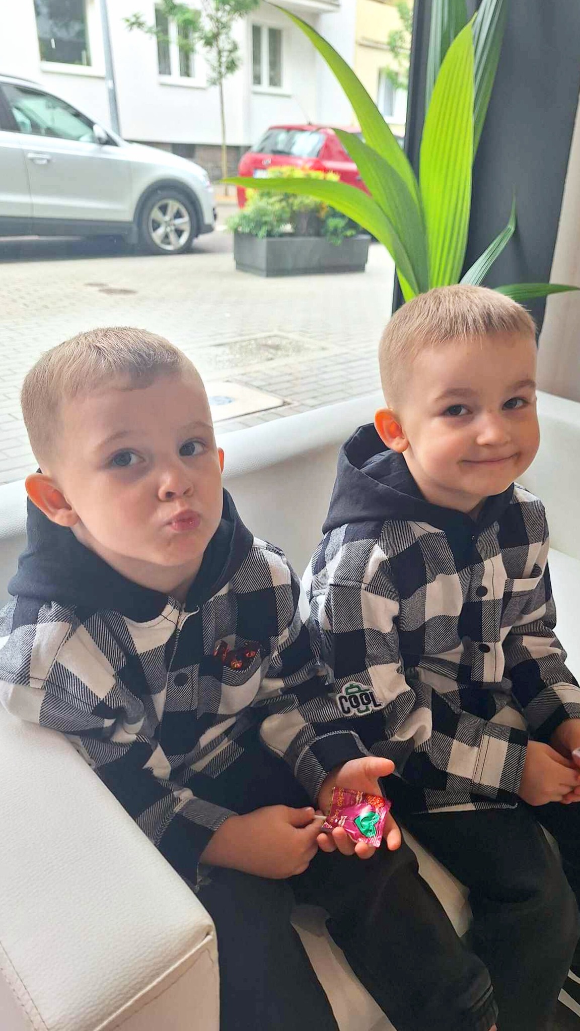 Dwaj mali chłopcy z krótkimi fryzurami w identycznych koszulach w kratę siedzą obok siebie na białej kanapie, jeden trzyma cukierek w dłoni.