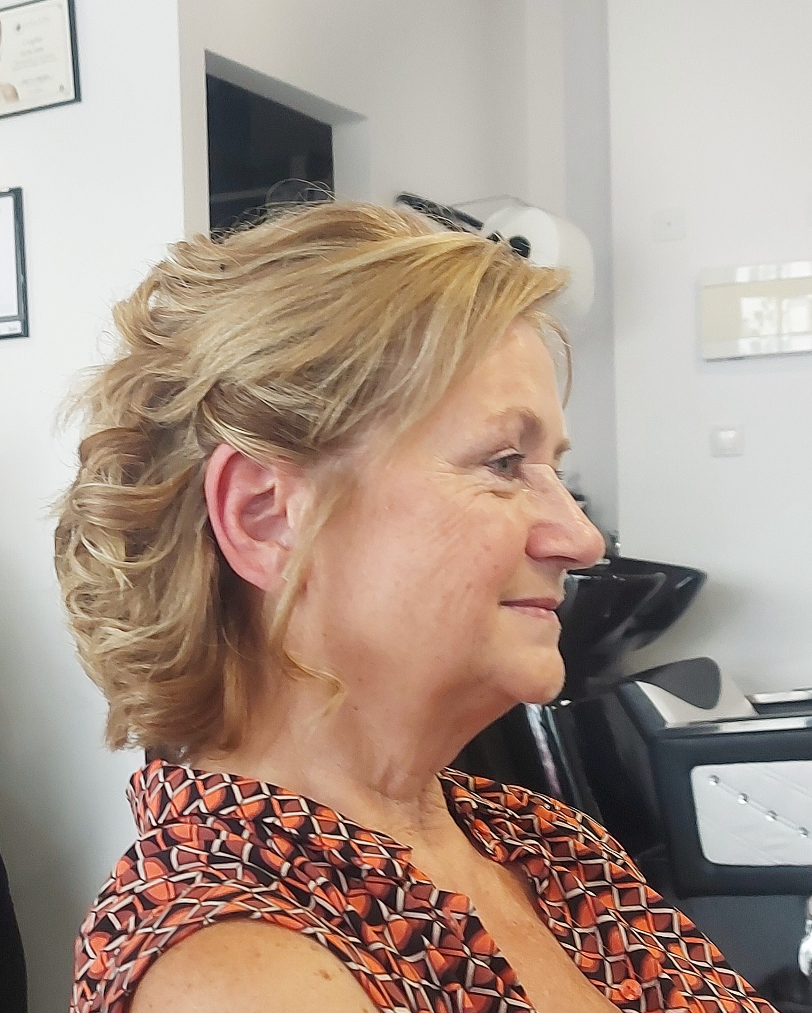 Profil kobiety z upiętymi, blond włosami w salonie, widoczna umywalka fryzjerska w tle.