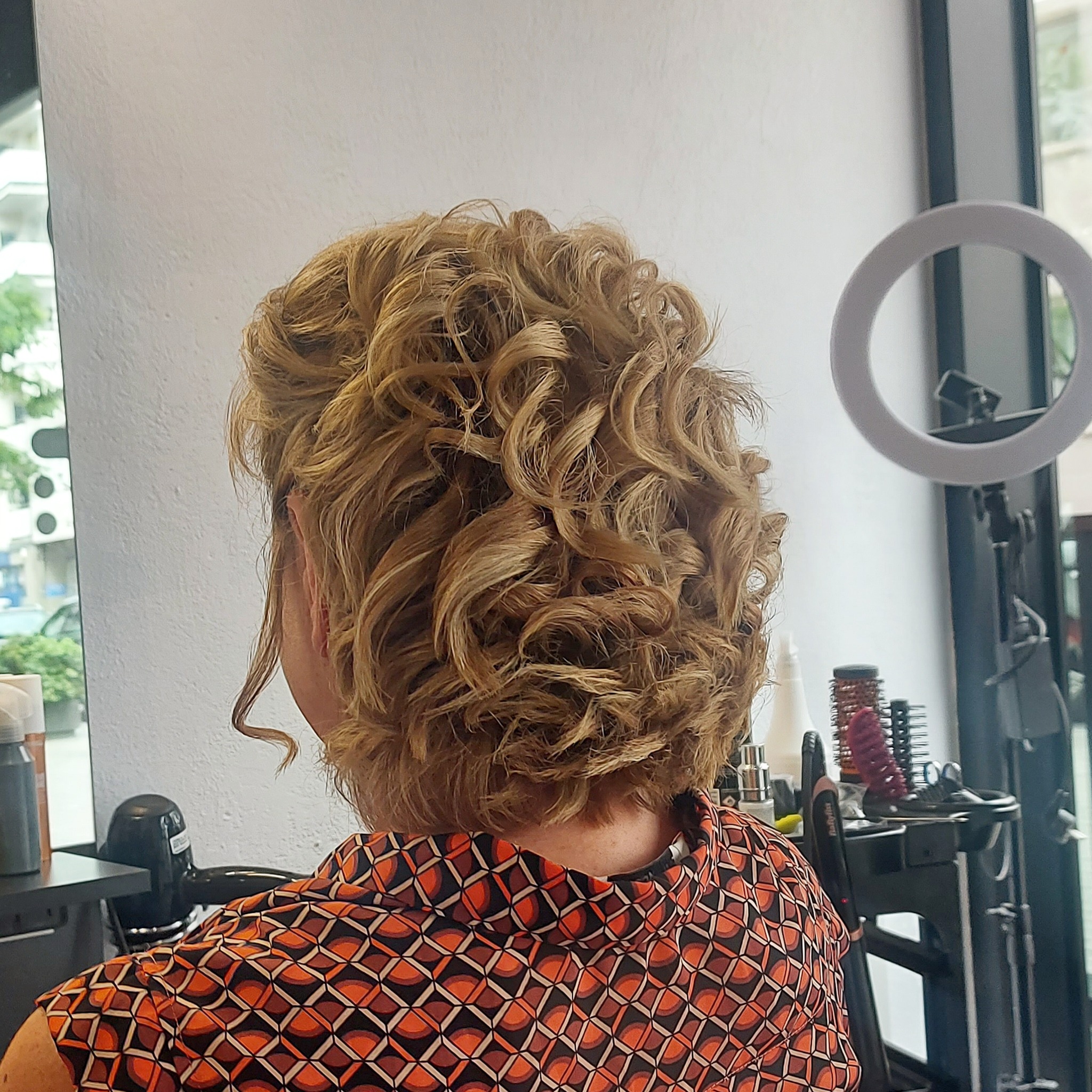 Upięcie z loków w odcieniach blondu, widok z tyłu na kark kobiety w pomarańczowej koszuli we wzory, w tle stanowisko pracy z narzędziami i ringiem doświetlającym.