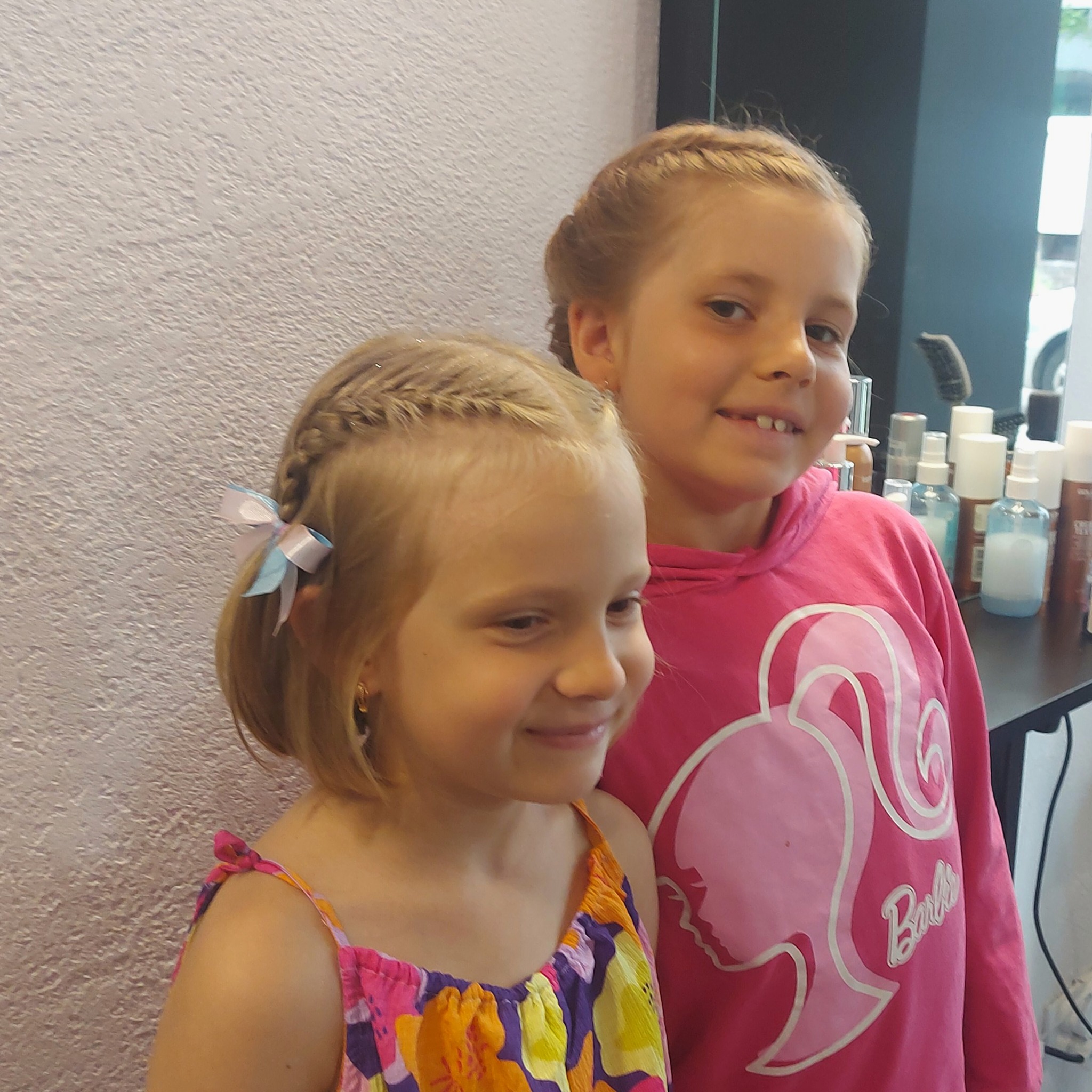 Dwie uśmiechnięte dziewczynki po wykonaniu fryzur z warkoczami, jedna z kokardką we włosach, w tle widoczne kosmetyki i akcesoria.