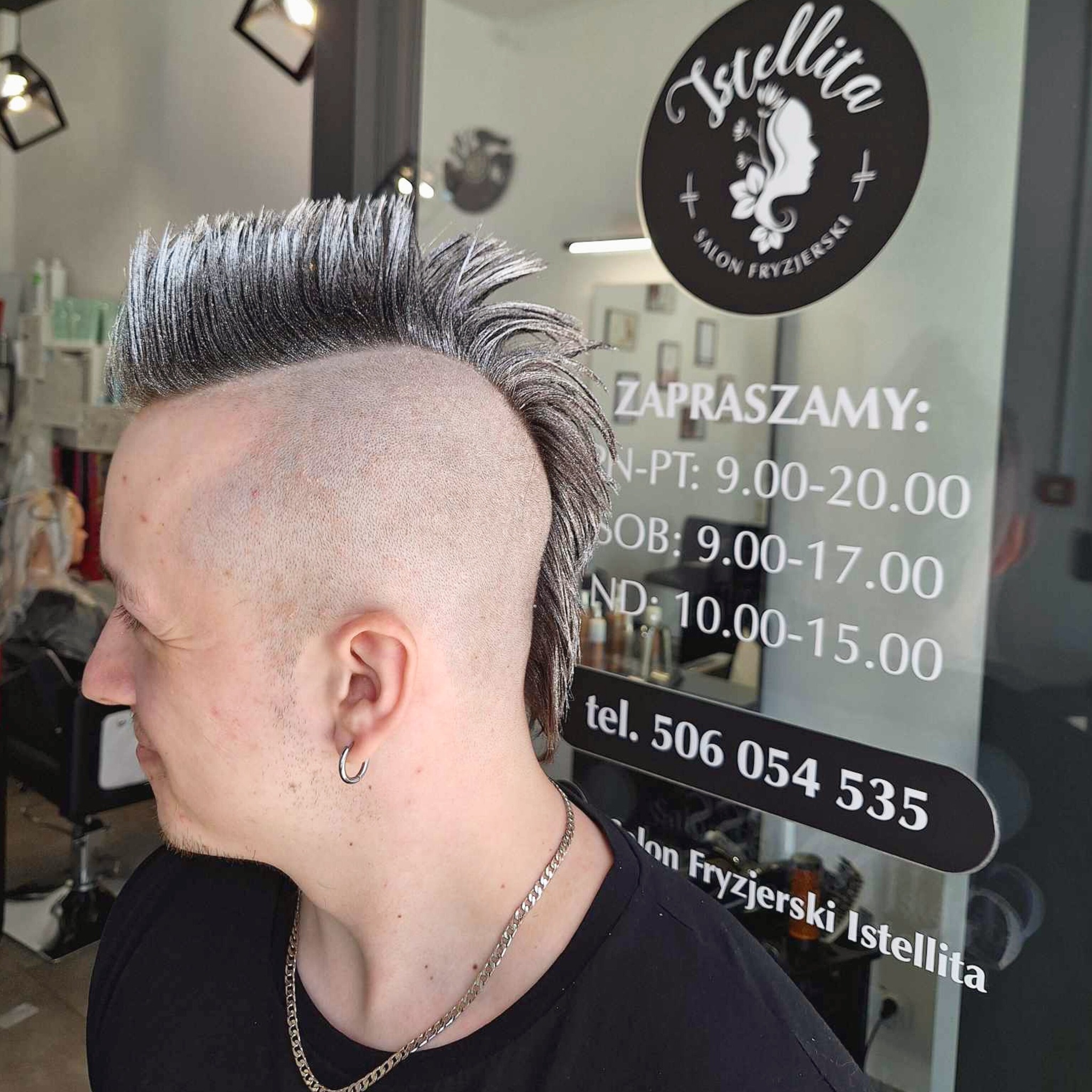 Mężczyzna z irokezem w odcieniach szarości, widoczny z profilu na tle witryny salonu fryzjerskiego Istellita z godzinami otwarcia i numerem telefonu.