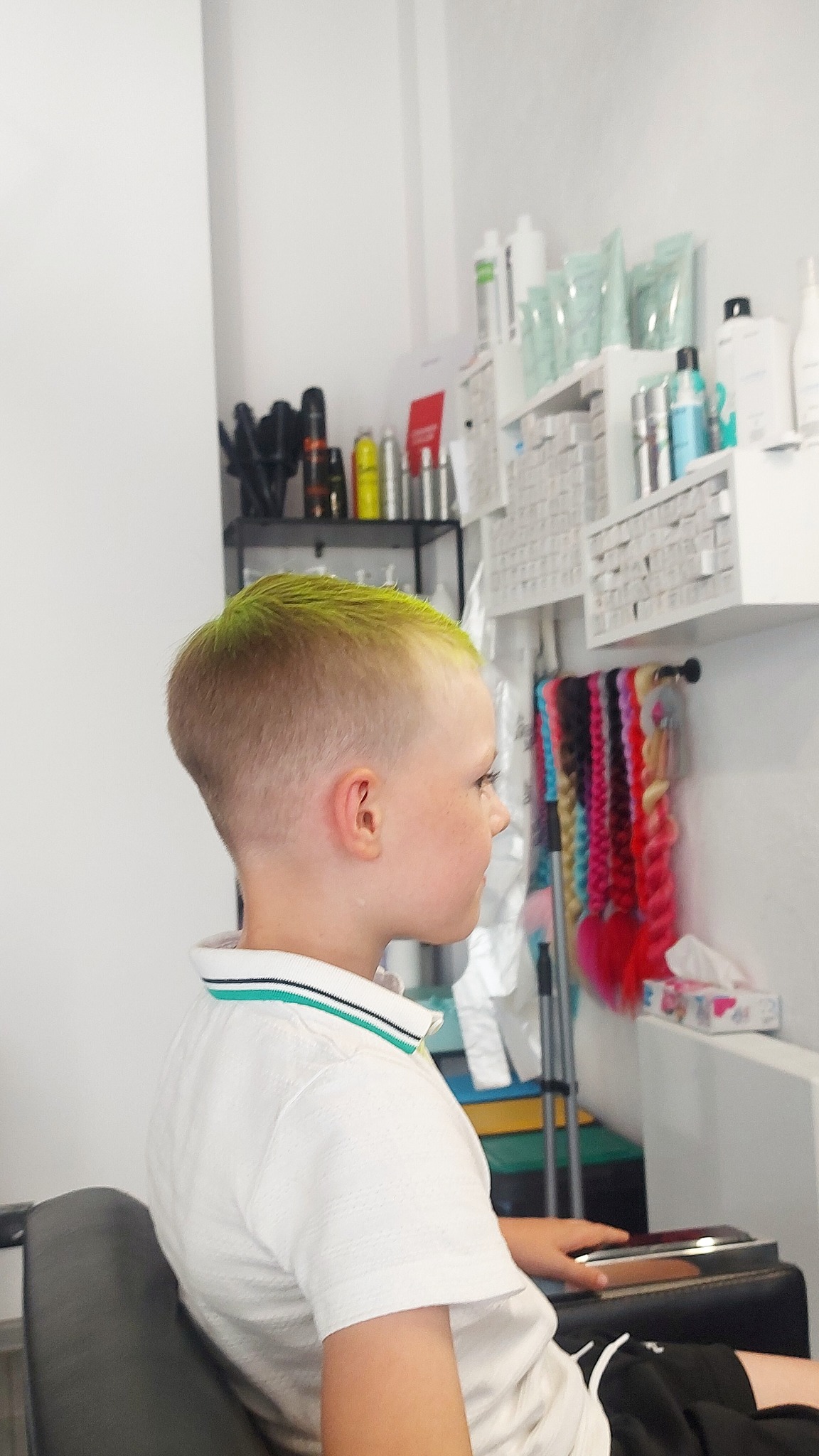 Profil chłopca z krótkimi, blond włosami z zielonymi końcówkami, siedzącego na fotelu w salonie, w tle półki z kosmetykami i kolorowe warkocze.