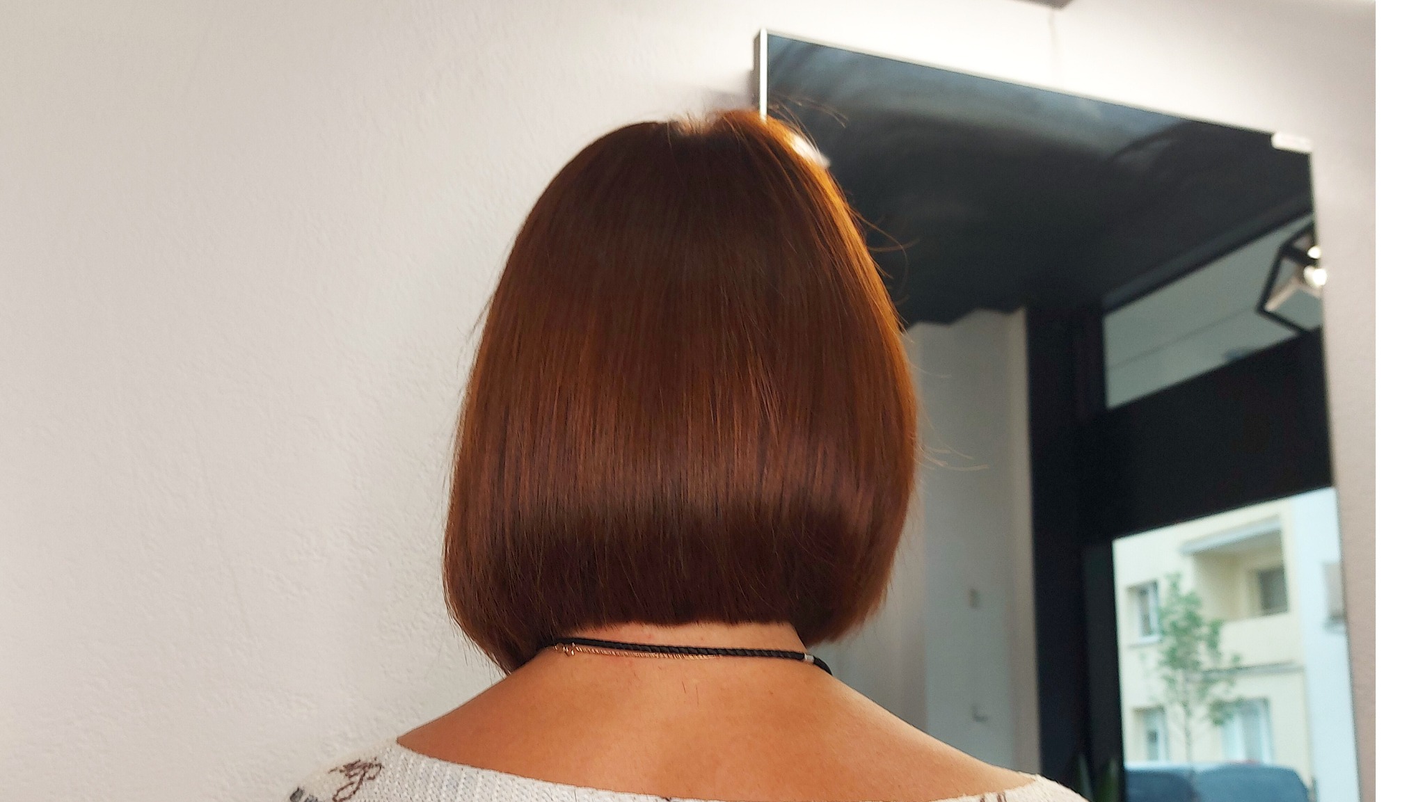 Krótkie, brązowe włosy w stylu bob, widok z tyłu, z delikatną biżuterią na szyi i odbiciem w lustrze wnętrza salonu.