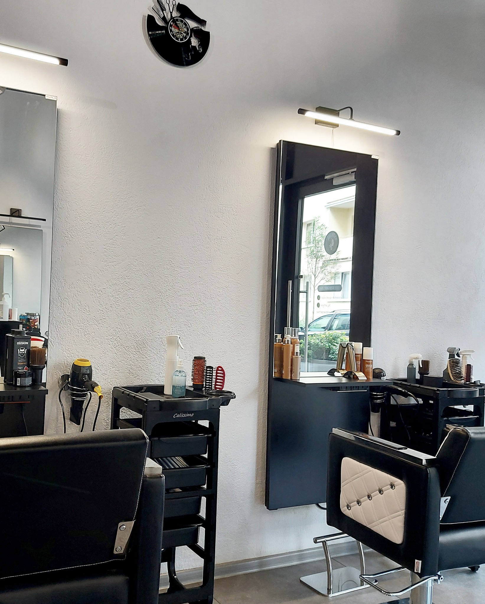 Wnętrze salonu, z czarnymi fotelami z pikowanymi oparciami, w tle wózki z akcesoriami i lustra podświetlone lampami oraz zegar z motywem fryzjerskim na ścianie.