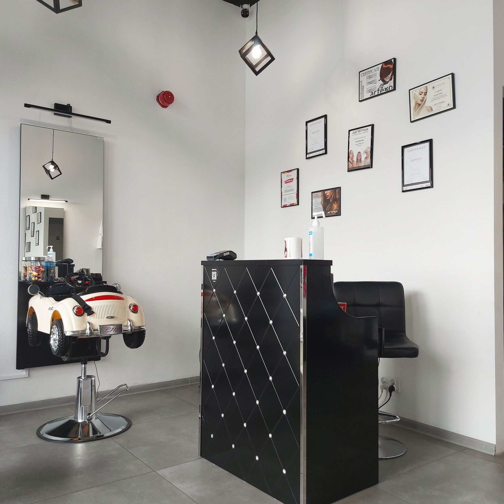 Wnętrze salonu z recepcją i krzesłem fryzjerskim dla dzieci w kształcie samochodu retro, na ścianie wiszą certyfikaty.