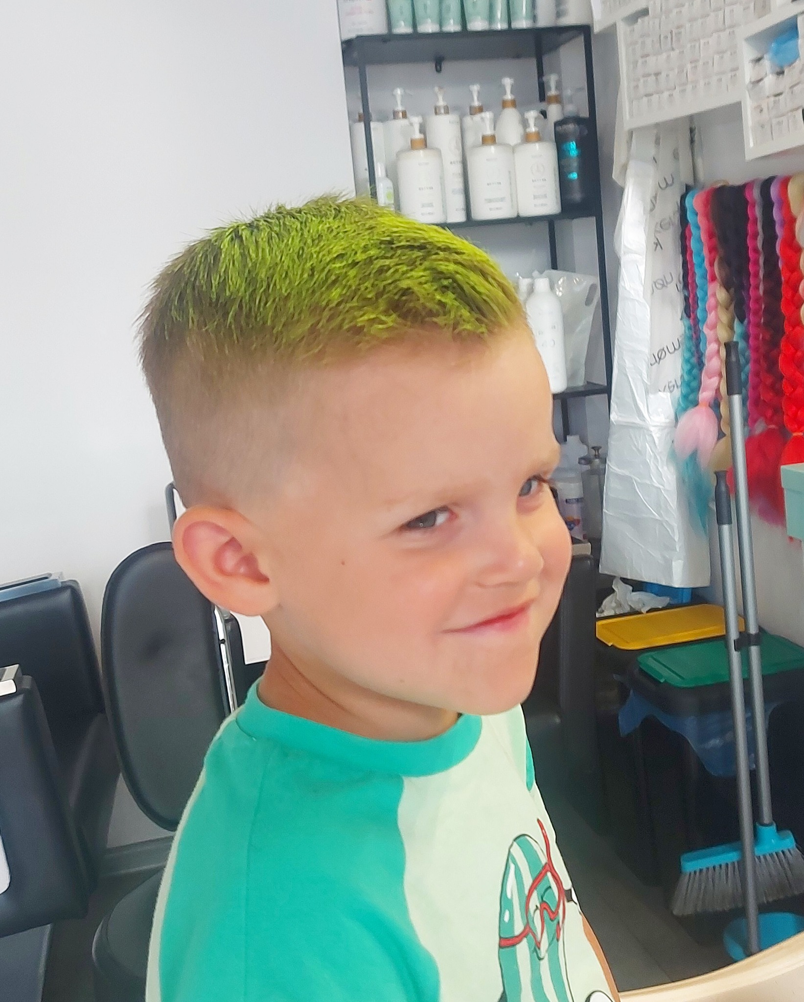 Chłopiec z krótką fryzurą i jaskrawo zielonymi włosami, uśmiechający się w salonie, na półce widoczne kosmetyki i kolorowe warkocze.