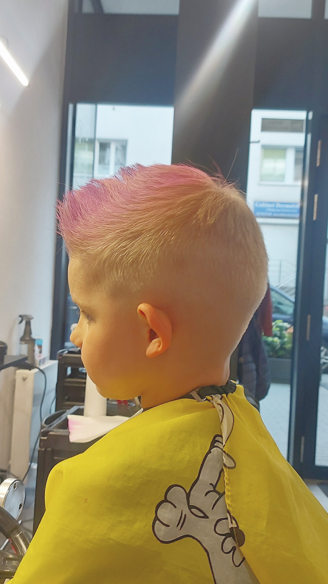 Chłopiec z krótką, blond fryzurą i różowymi końcówkami, w żółtym pelerynie z nadrukiem, widok z profilu w salonie.