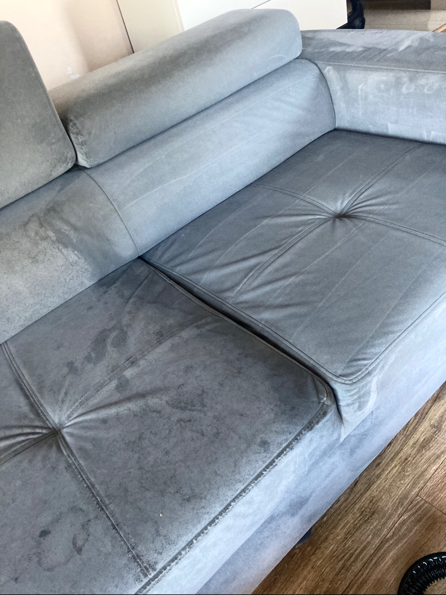 Szara, narożna kanapa z widocznymi plamami przed czyszczeniem, z fragmentem odkurzacza piorącego w rogu.