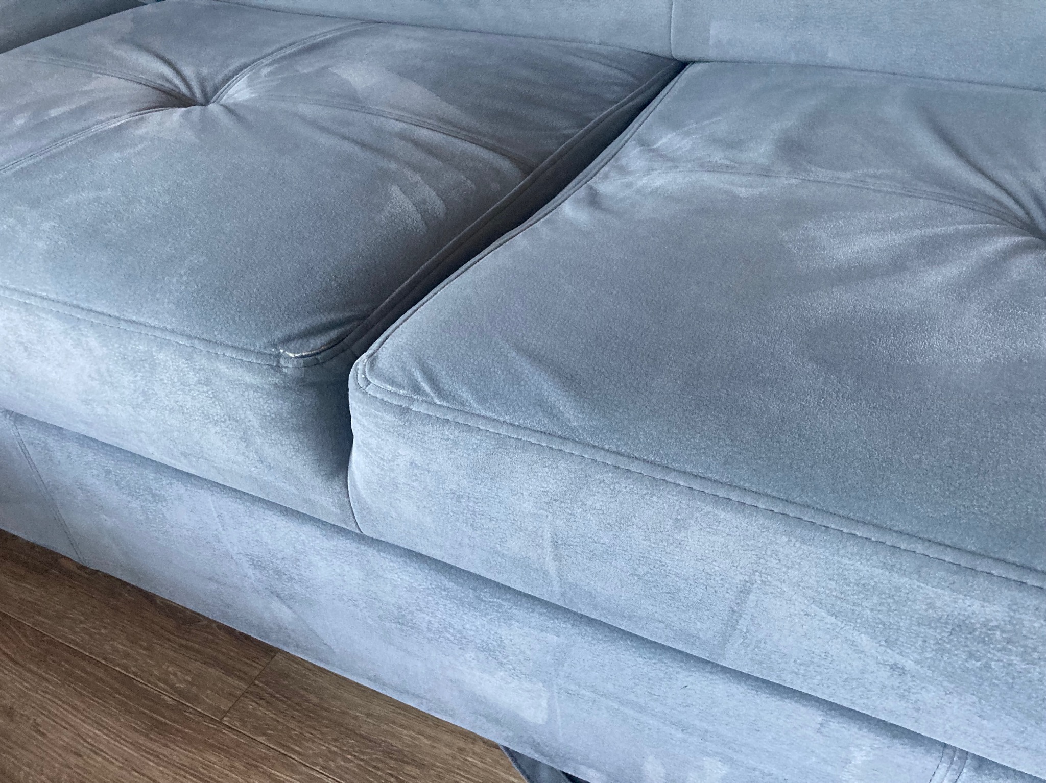 Jasnoszara, welurowa kanapa modułowa z widocznymi plamami po czyszczeniu na siedzisku, ustawiona na panelach podłogowych.