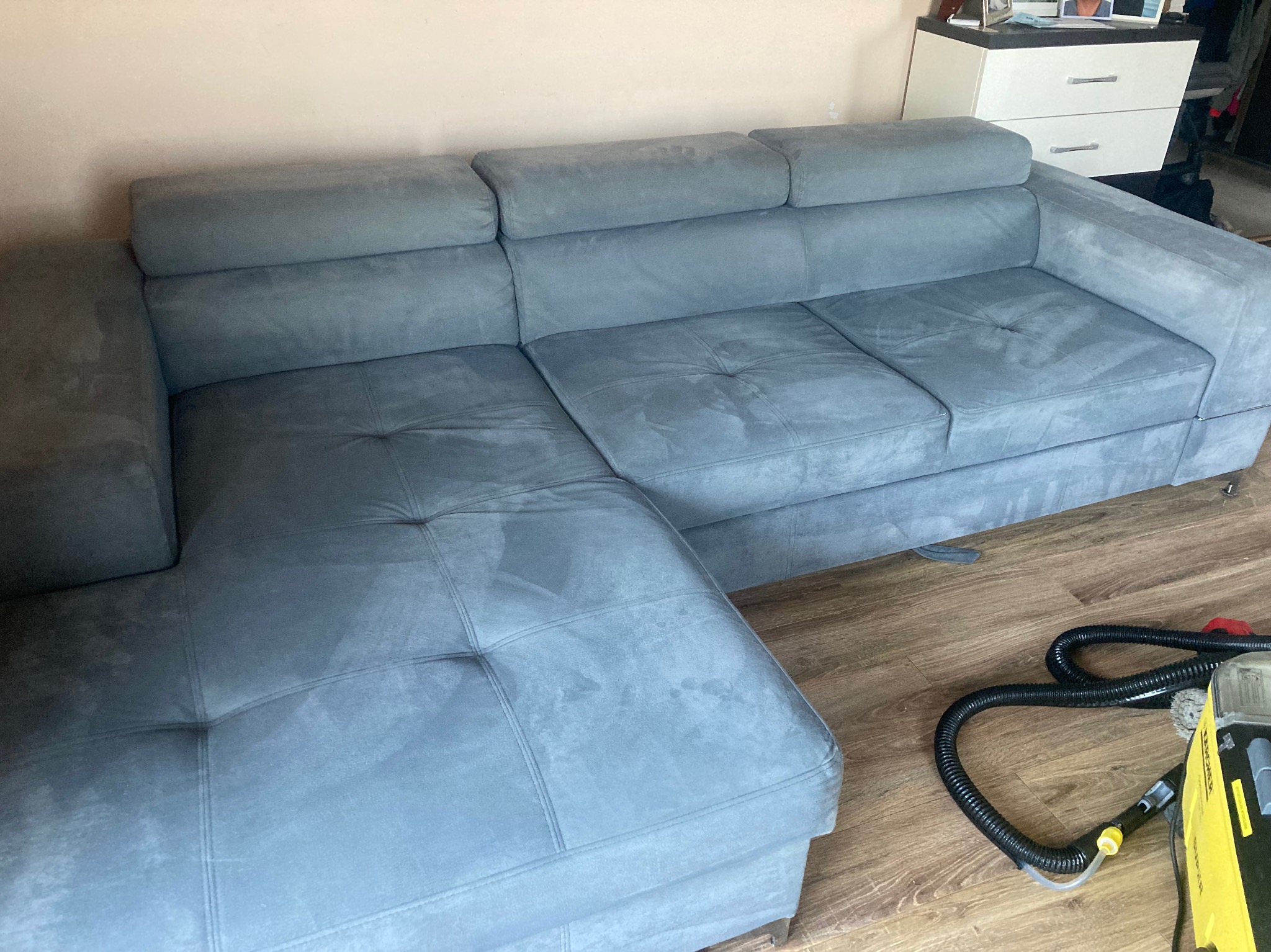 Szara narożna kanapa z widocznymi śladami czyszczenia na tapicerce, obok leży wąż od odkurzacza piorącego na drewnianej podłodze.