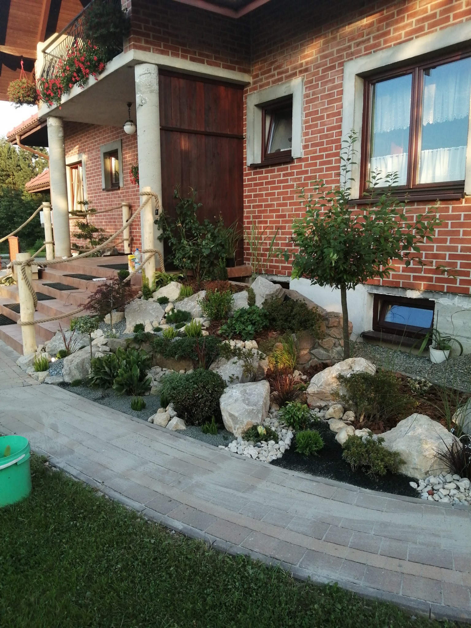 Ogród skalny z różnorodną roślinnością u podnóża ceglanego domu, z widocznym chodnikiem z kostki brukowej i schodami z drewnianymi balustradami.