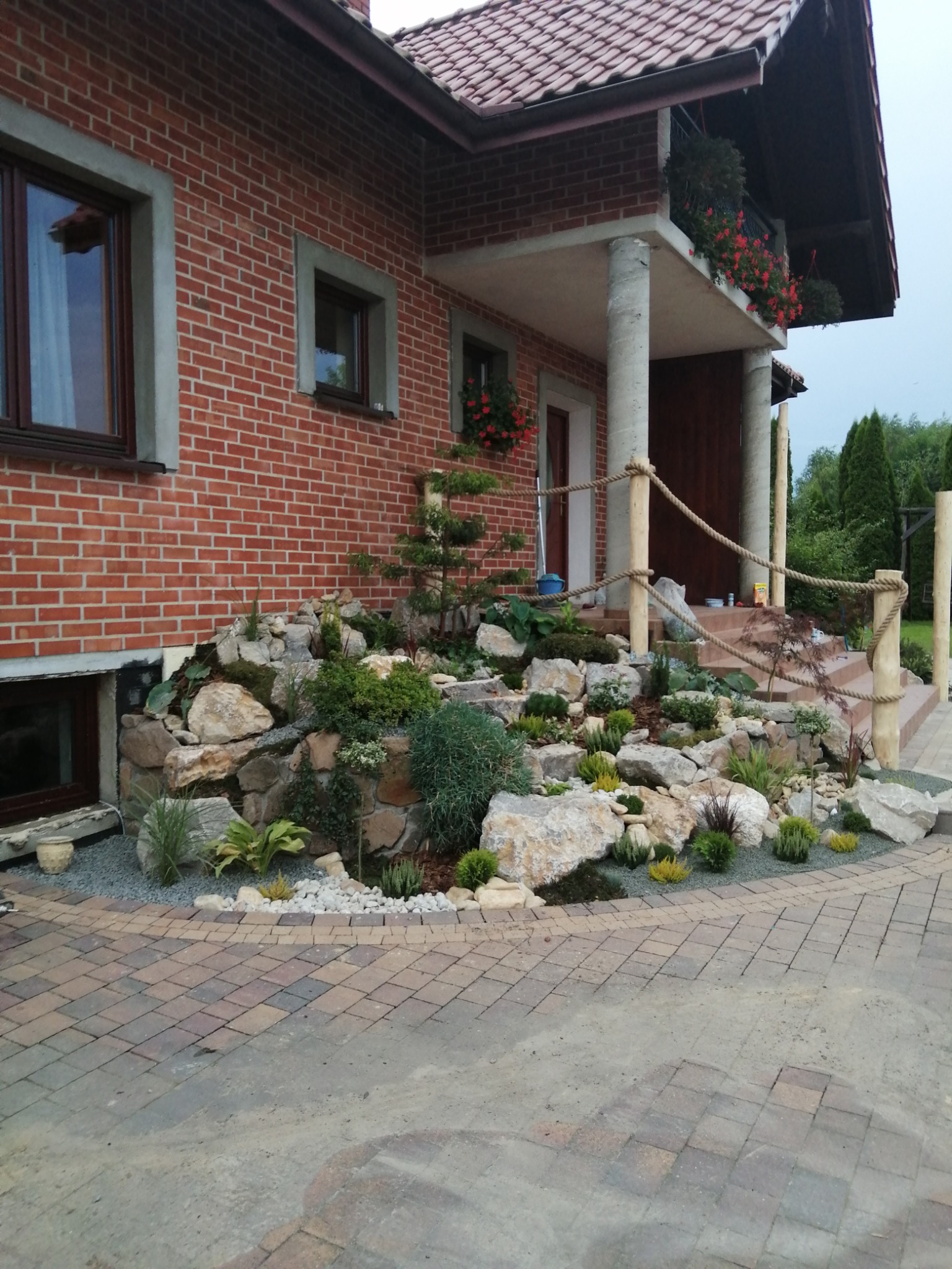 Ogród skalny przed ceglanym domem z drewnianymi schodami i balustradą z liny, z różnorodną roślinnością i kamieniami różnej wielkości