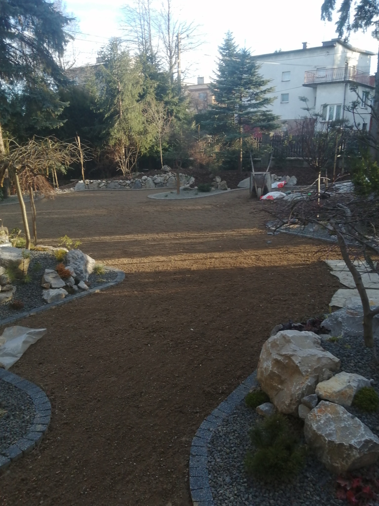 Przygotowany teren pod trawnik z obrzeżami z kostki granitowej, kamieniami ozdobnymi i młodymi drzewkami, widoczne worki z ziemią, w tle zabudowa mieszkalna.